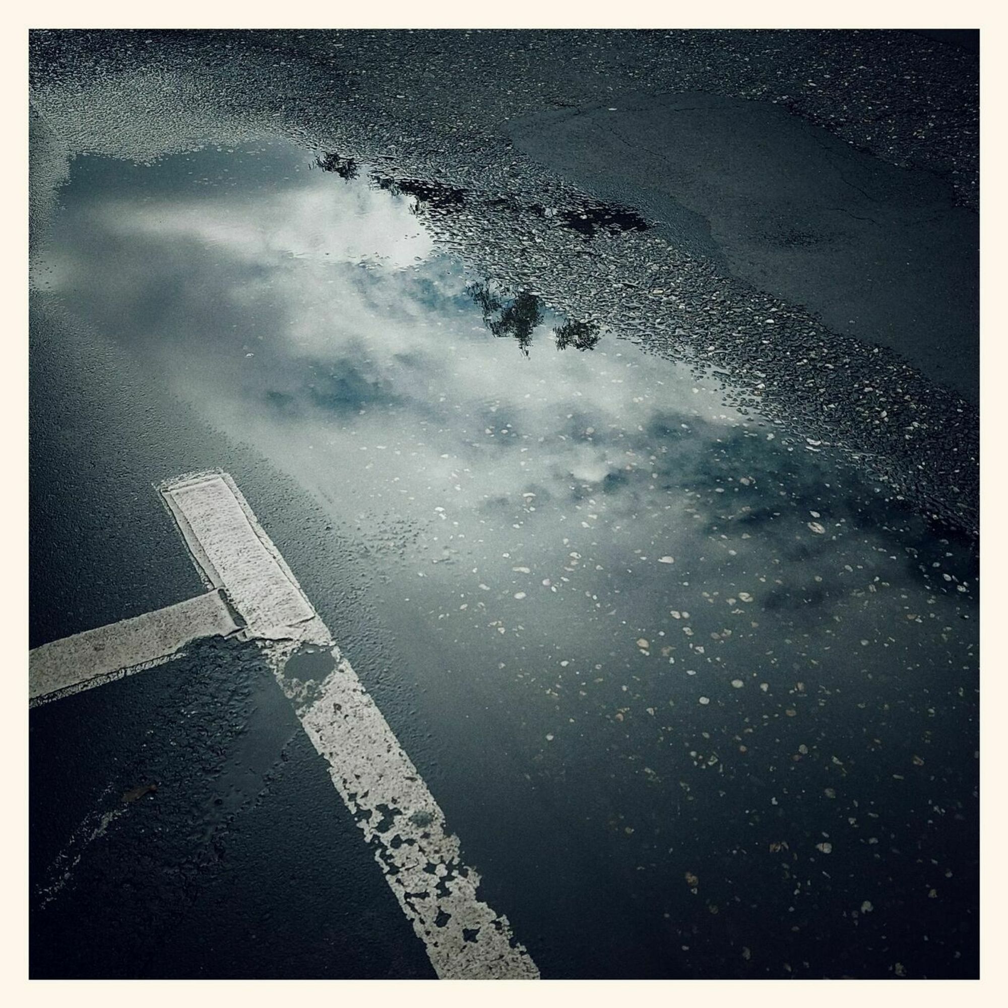 Reflections of clouds and blue sky on a flat puddle of water. Guiding lines of a parking lot left of it.