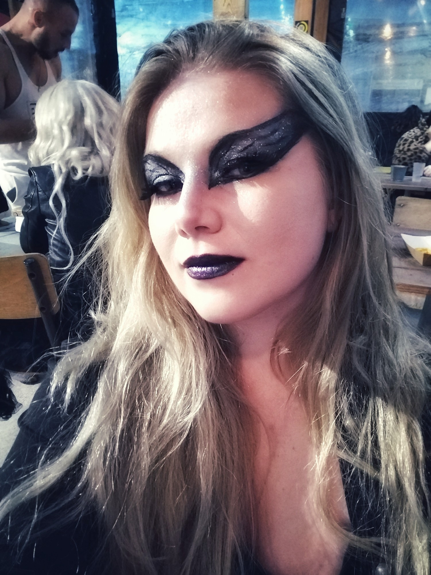 3/4 face of a blond woman with long hair wearing a black swan full of glittermake-up and black lips