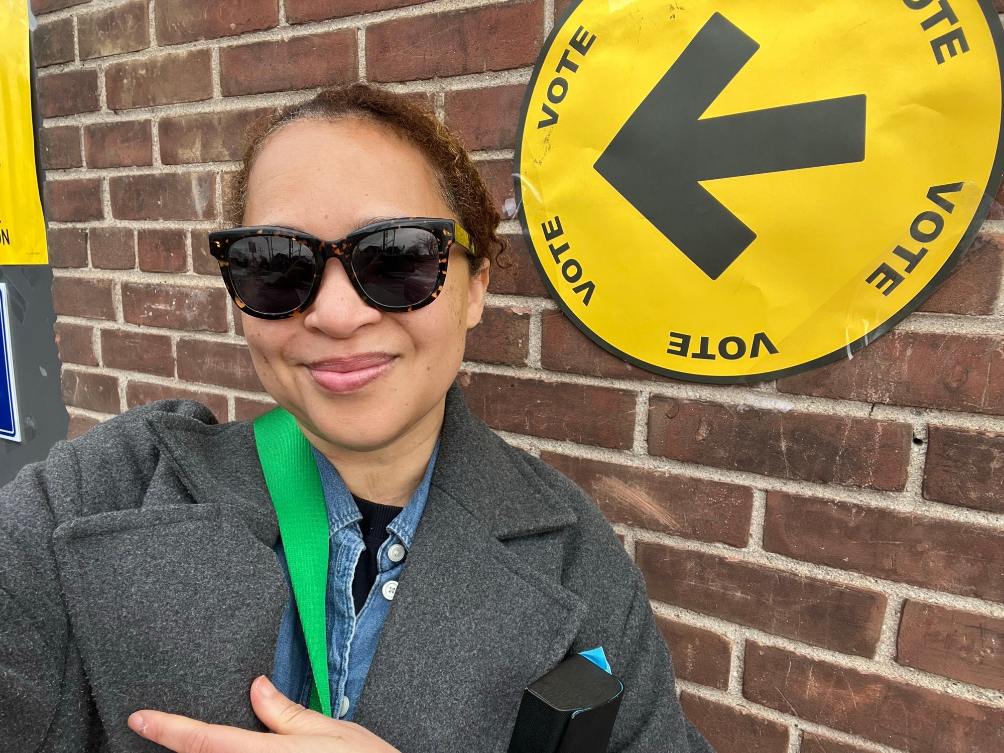 Picture of Nam Kiwanuka with sunglasses in outside of an election office. Behind them is a sign with an arrow pointing the direction to vote.