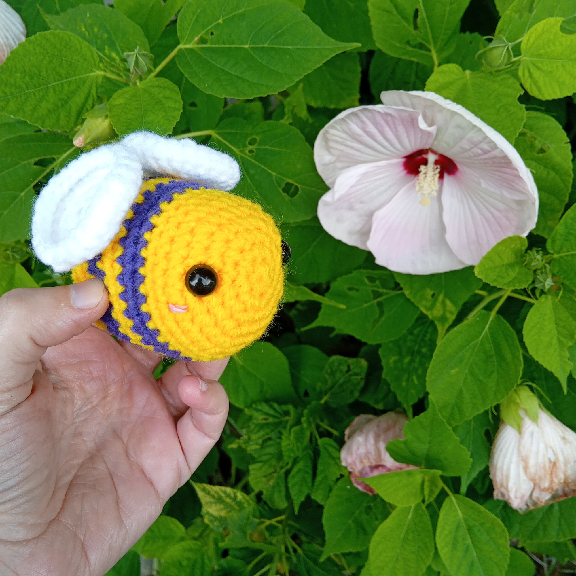 Abeille au crochet. Elle est jaune, avec des rayures violettes, des ailes blanches et des yeux noirs. Elle est devant un hibiscus avec une fleur rose pâle.