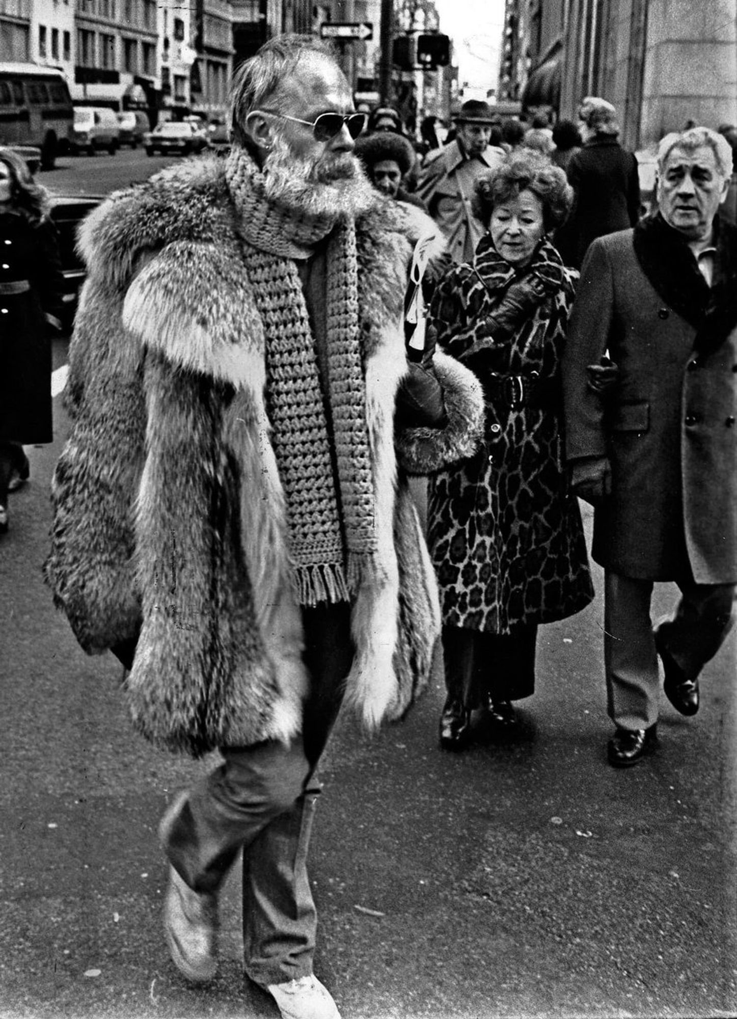 A B&W photo from the 1970s of illustrator Edward Gorey, looking tall, burly and authoritative as he wears a huge furry coat (possibly a raccoon coat) and sunglasses on a NYC street, with other well-dressed New Yorkers watching him.