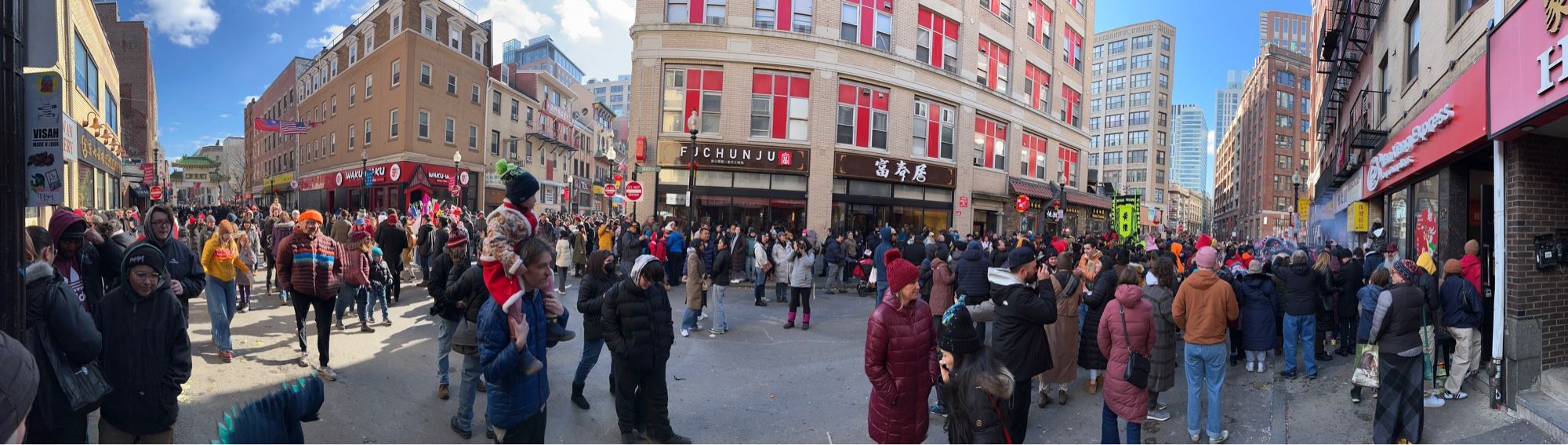 Panoramic picture of a Chinese lunar New Year celebration with a large crowd, possibly, in an area with Chinese signage, suggesting a Chinatown Boston. People are dressed in winter clothing, suggesting cold weather.