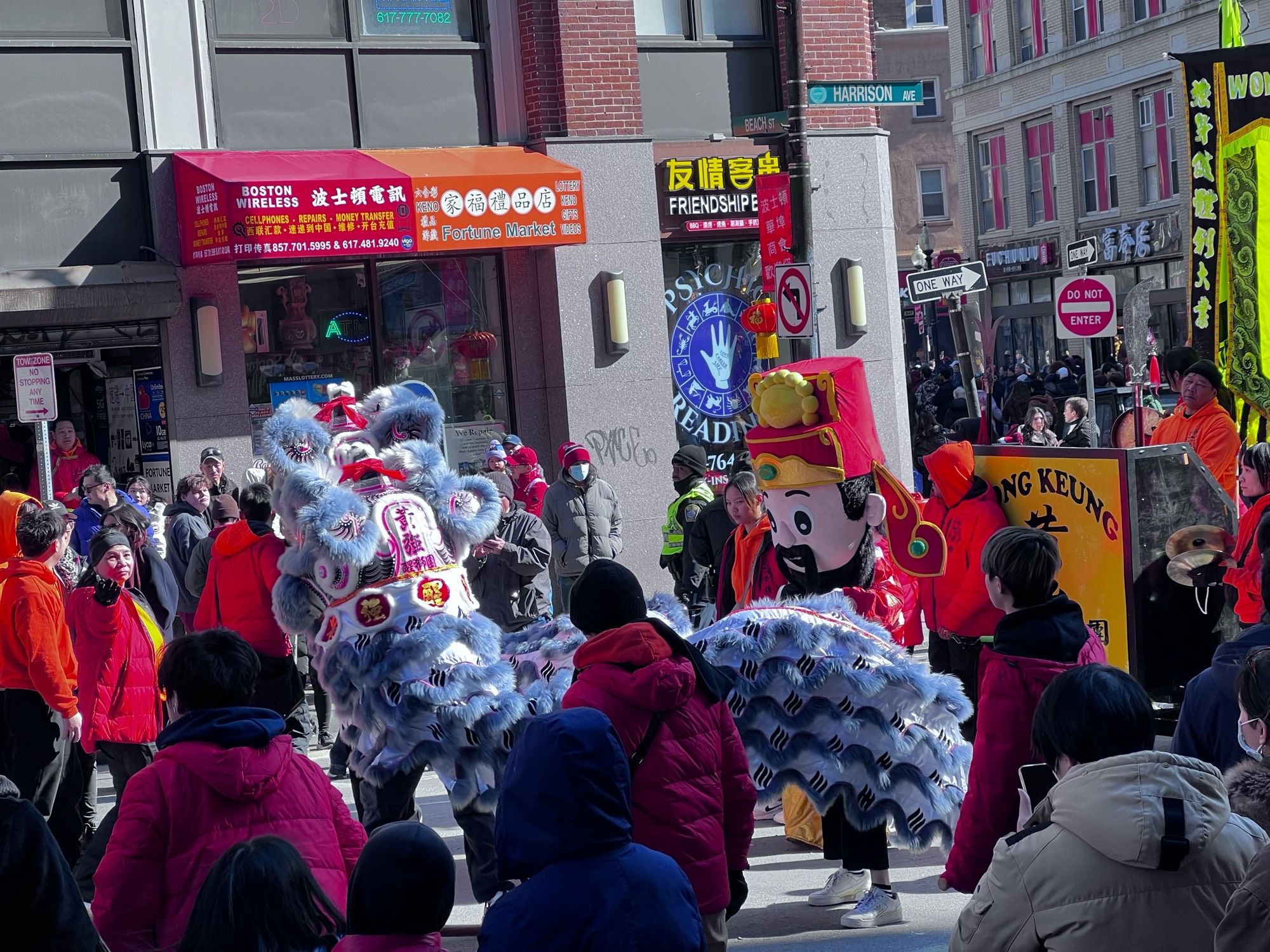 A lively street scene with people watching a Chinese lion dance performance during lunar new year 2024 in Boston. There is a large costumed figure resembling a historical Chinese character, various banners with Chinese writing, and spectators in winter clothing.