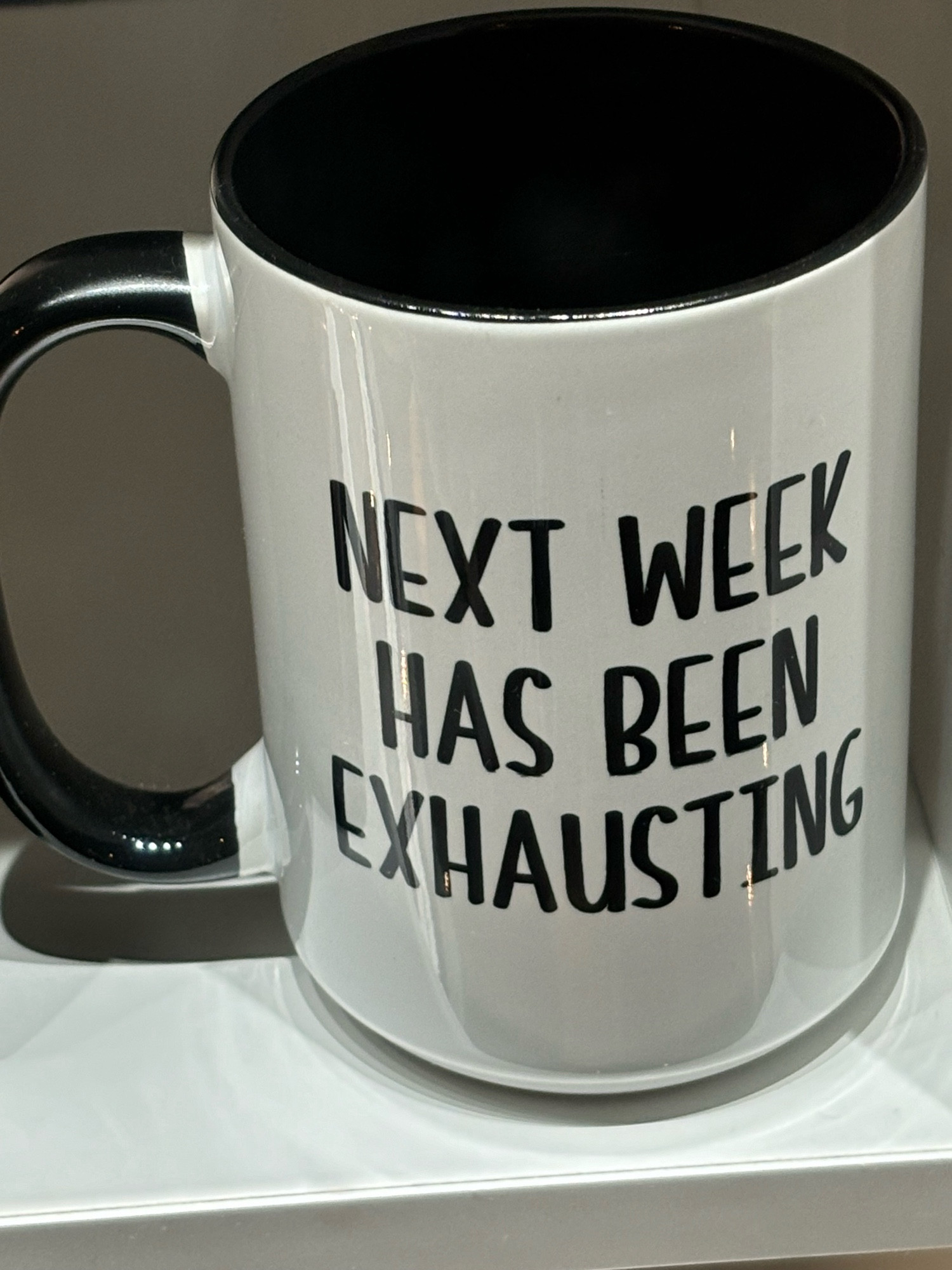 White coffee mug with black handle and printed text reading “Next Week Has Been Exhausting”