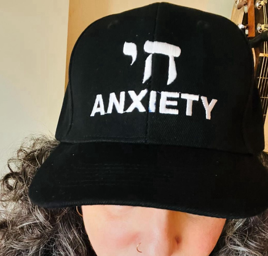 Black baseball cap with printing: Hebrew letters forming the word “chai” (meaning life) and the word “anxiety”