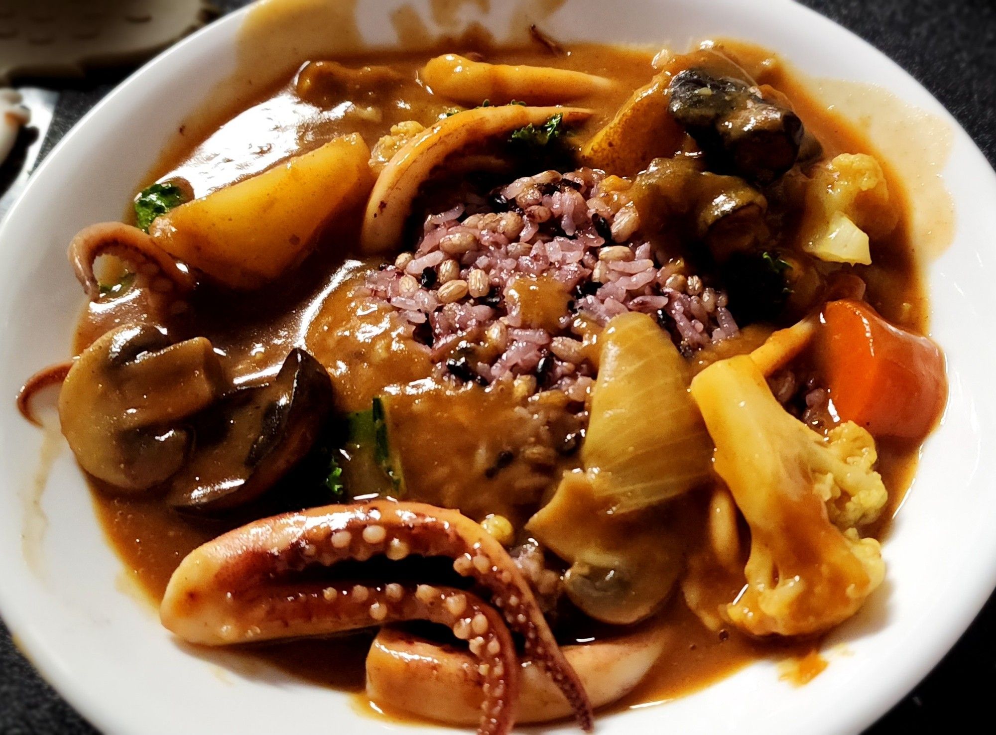 A shallow bowl of curry served over a mound of purple rice. The curry has cauliflower, squid tentacles, carrots, white and woodear mushrooms, onion, and potatoes.