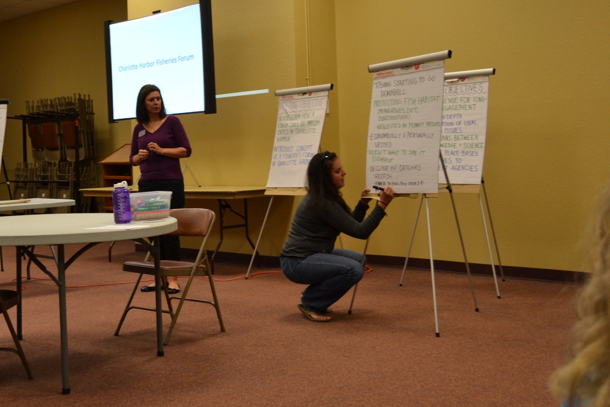 Woman crouches on the ground in a carpeted room in front of a flip chart, writing on it with a marker. Another woman stands next to her in front of a projector screen.