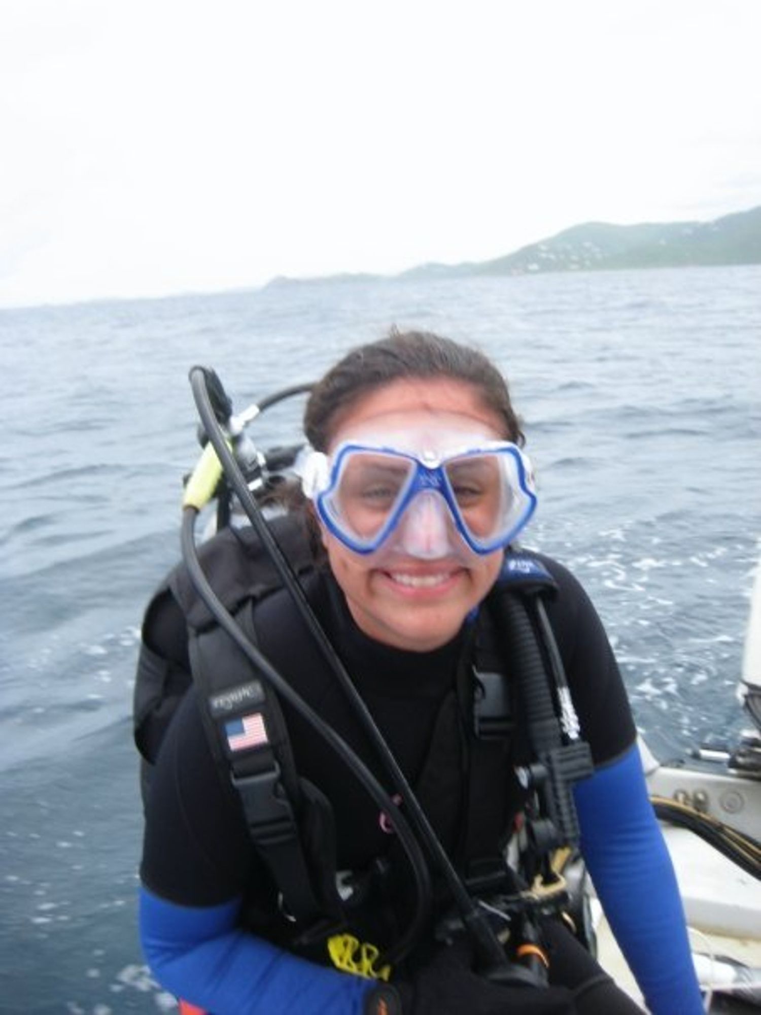 Young woman sitting on the edge of a boat in scuba gear with facemask on, smiling at the camera. Behind her is water and in the distance an island shore can be seen.