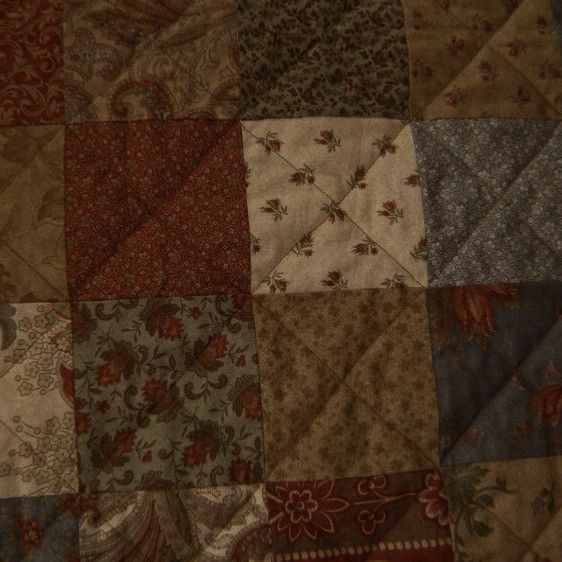 Zoomed in quilt with many stretched together squares of differing patterns of cloth. Many of which are flower designs in a warm color