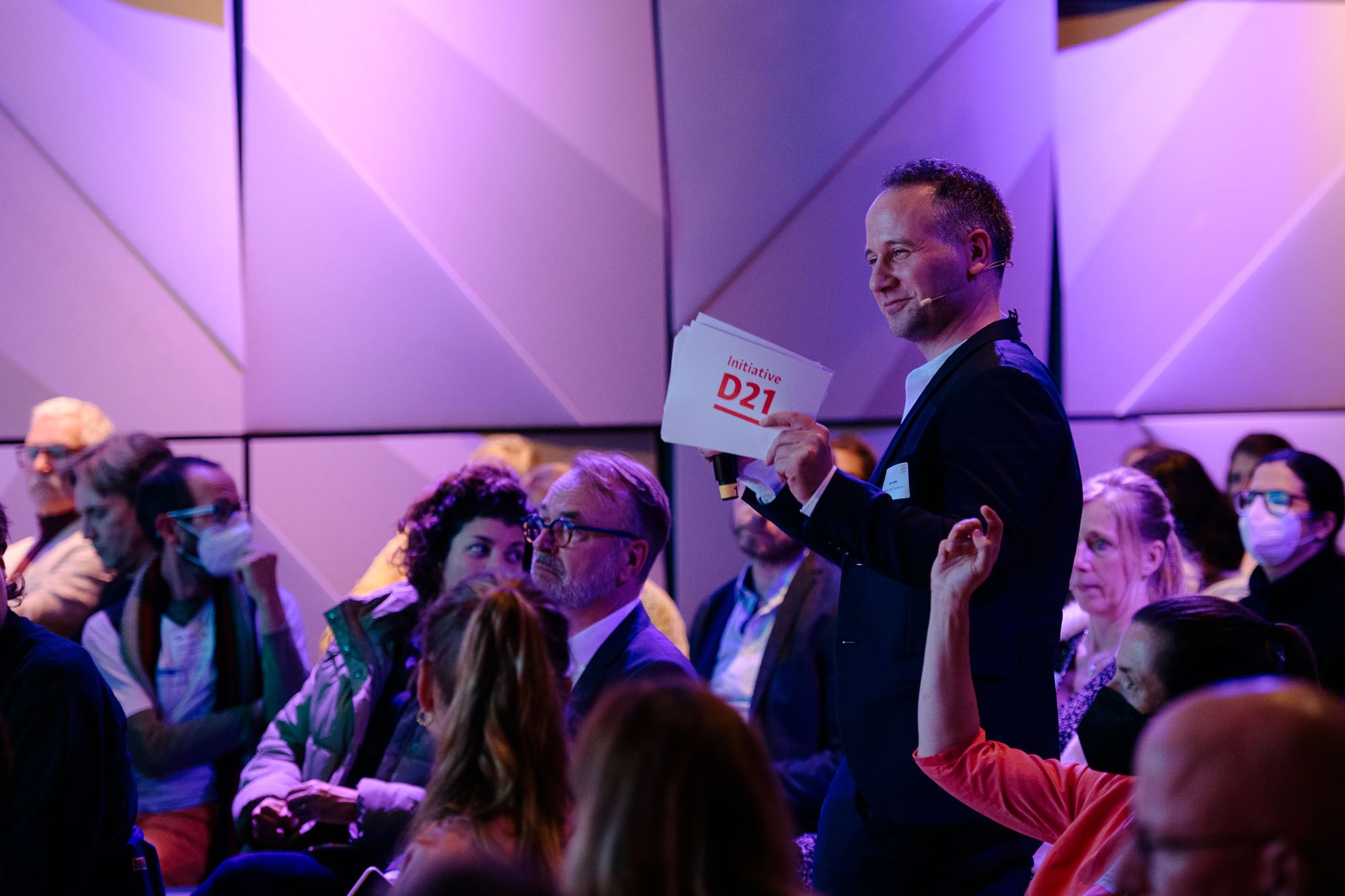 Foto von Timm Lutter inmitten des Publikums einer Veranstaltung. Er hat Moderationskarten mit D21-Logo in der Hand und gestikuliert.