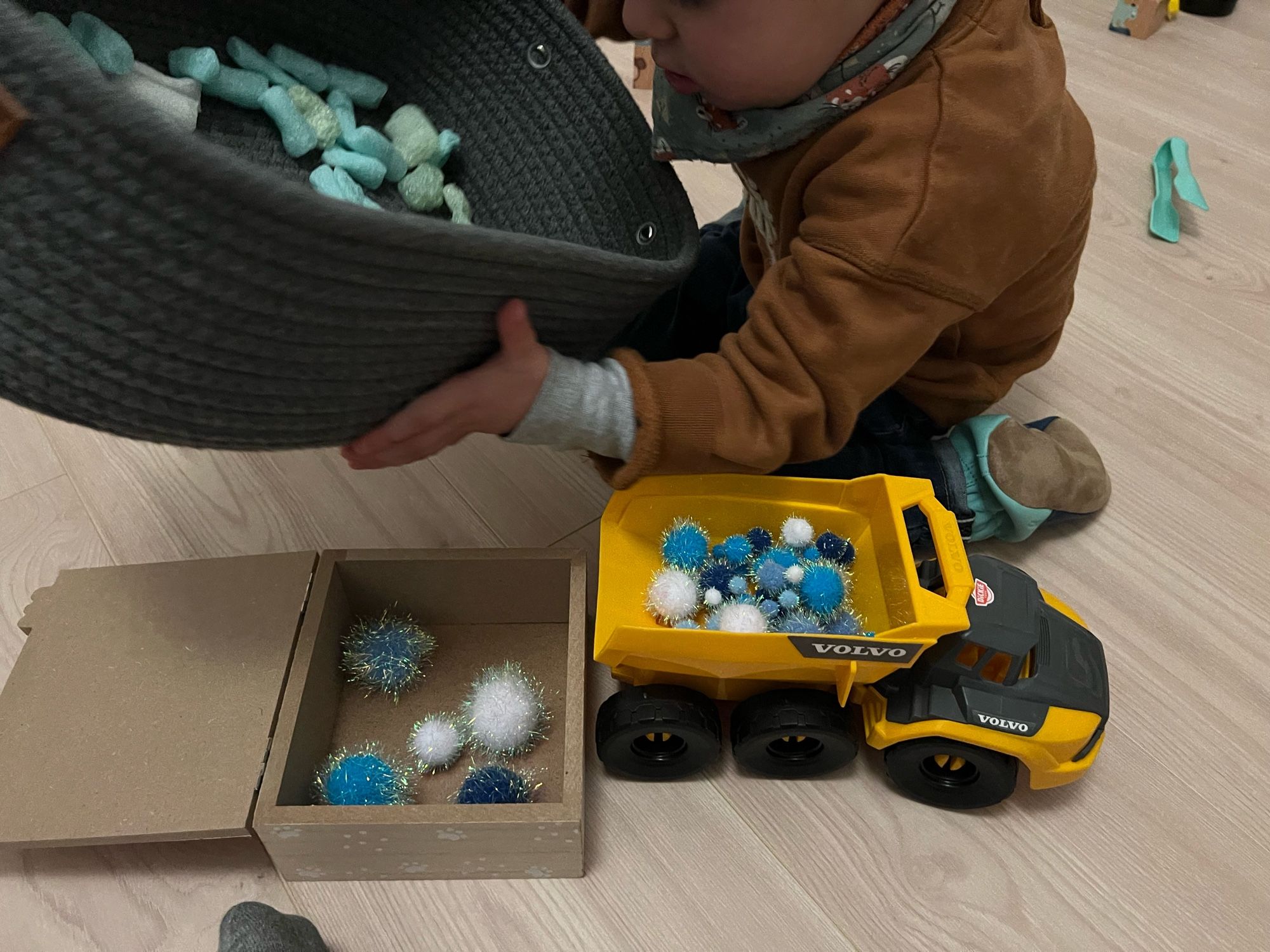 Activité transvasement de pompons de différentes tailles dans un camaïeu de bleus et blanc avec des filaments pailletés, dans un panier en tissu gris tenu par l’enfant, puis dans un jouet camion benne gris et jaune, puis dans une boîte en bois