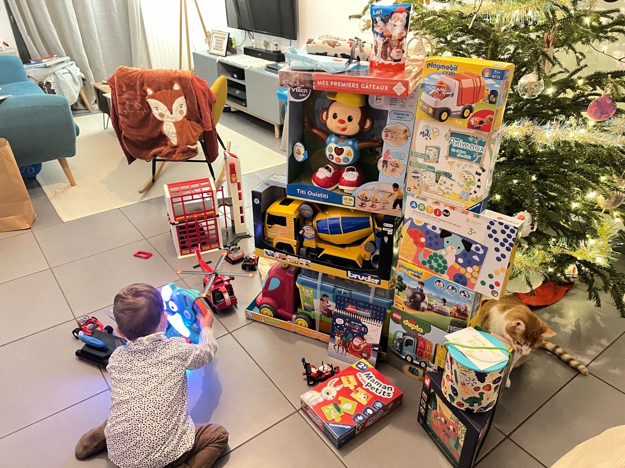 Le Mini jouant avec un de ses jouets (Solal le Narval) à côté d’un lot de Playmobil pompiers (caserne, hélico, bateau, les personnages et accessoires sont cachés), plus en avant se trouvent deux piles de jouets divers et variés d’environ 1m20 chacune, le tout devant le sapin. En bas à droite, Cléo le chat qui renifle et dépose ses phéromones sur un des jeux.