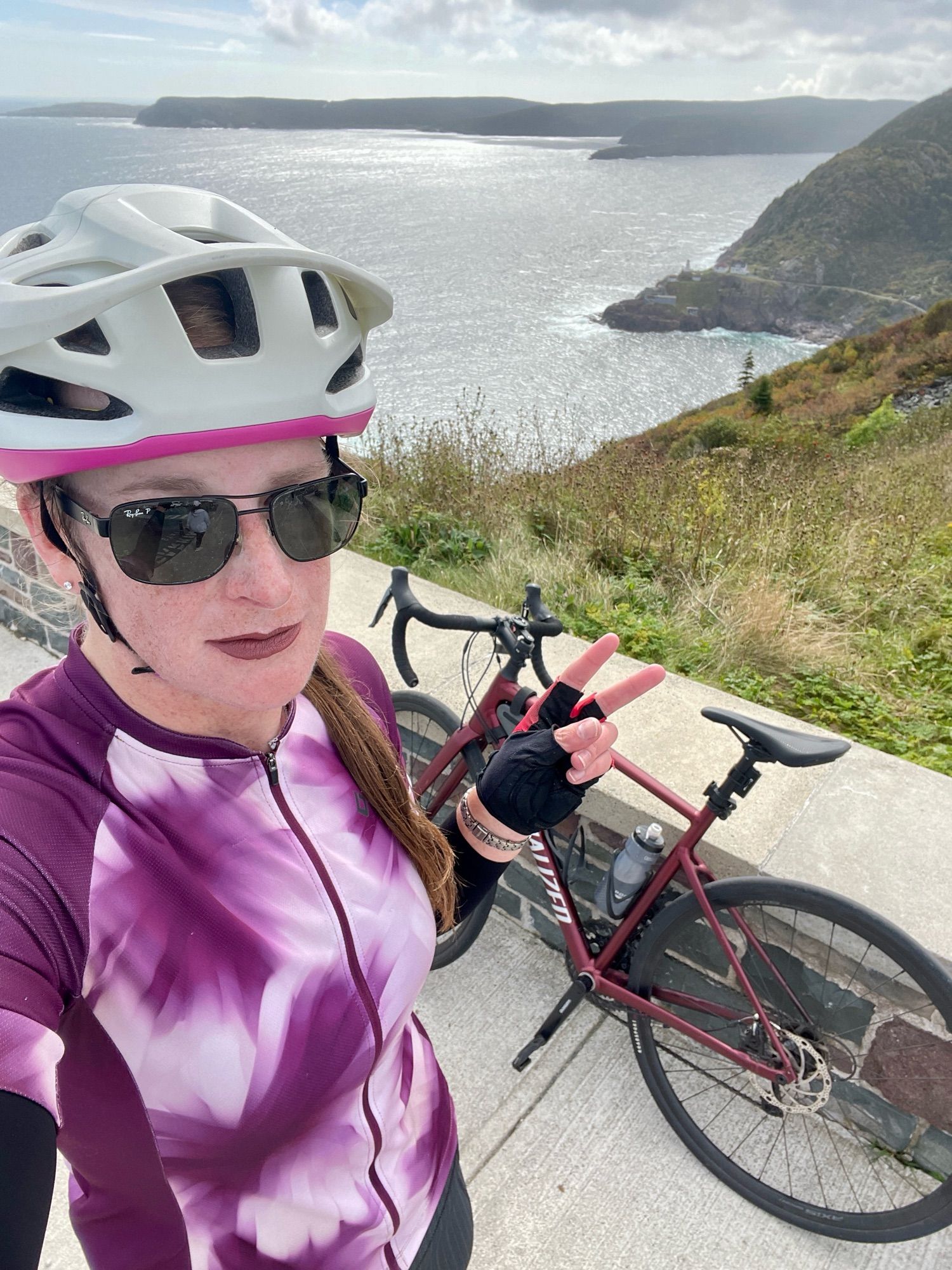 Cailín is standing on top of Signal Hill. Behind her is the Atlantic Ocean, Cape Spear, Fort Amherst, and her maroon Specialized Allez road bike. She’s wearing a white and fuchsia cycling helmet, black aviator-style sunglasses, mauve lipstick, a purple and white cycling jersey, and red and black cycling gloves.