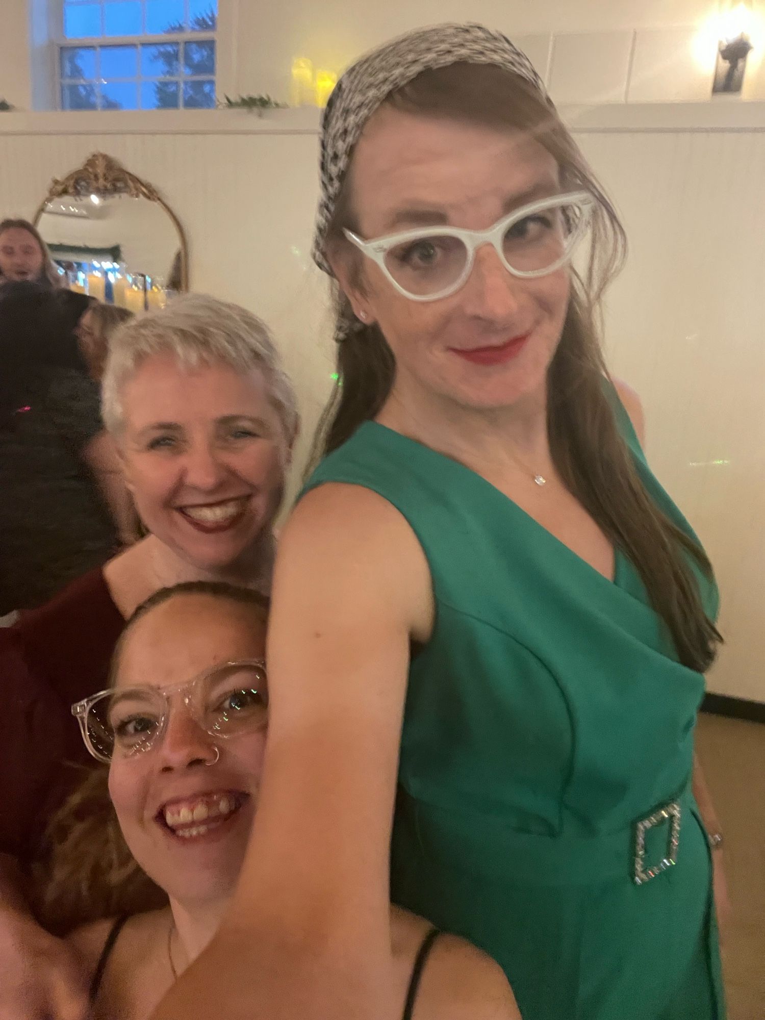 Cailín is taking a selfie with Eva and a mutual friend of theirs. Cailín is wearing a houndstooth bandanna, white cats eye glasses, red lipstick, and a green sleeveless jumpsuit. Eva is wearing burgundy lipstick and a burgundy dress. Their friend is wearing clear framed glasses and a strappy dress.