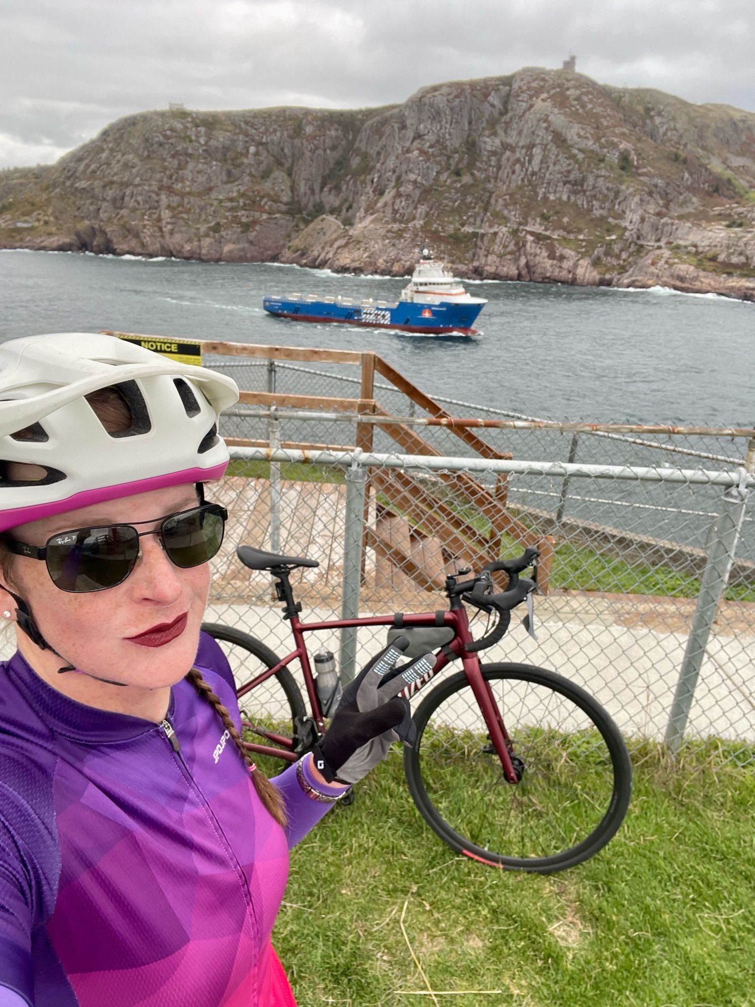 Cailín is standing at the end of the trail at Fort Amherst. Behind her is Cabot Tower on top of Signal Hill, a blue and white offshore supply vessel leaving St. John’s harbour via. the narrows, and her bike.