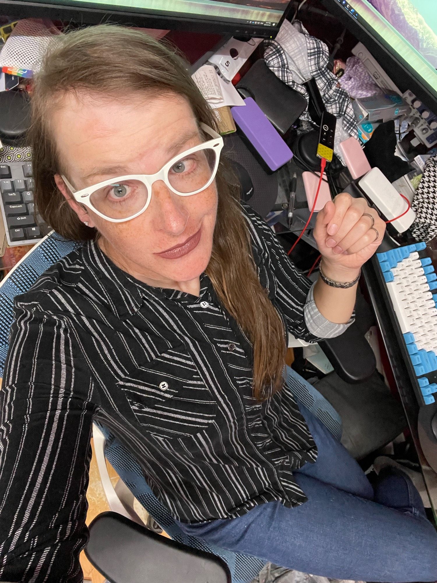 Cailín is sitting at her desk. She’s wearing white cats eye glasses, mauve lipstick, a black and white vertically striped blouse, and blue jeans