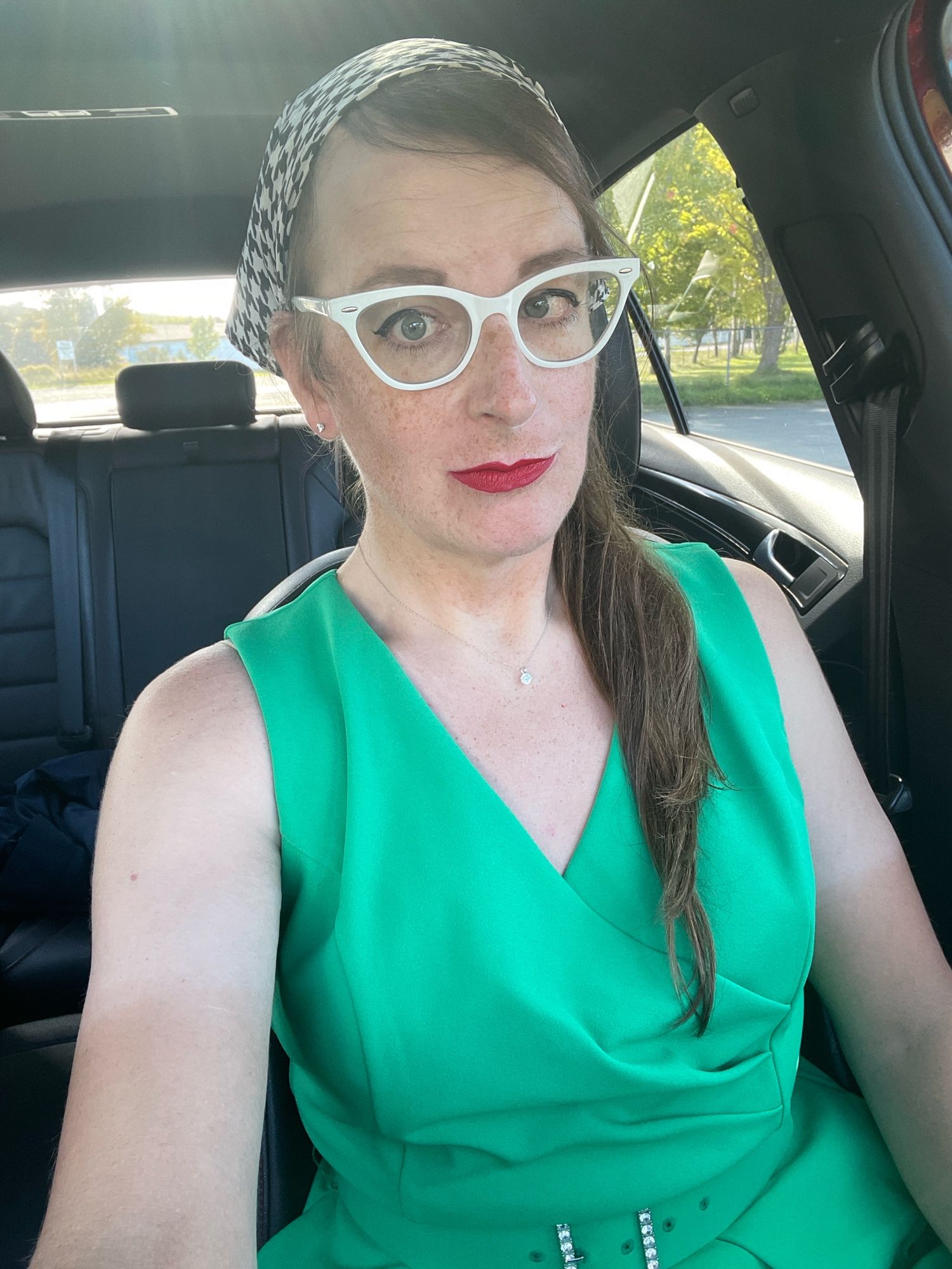 Cailín is sitting in her car. She’s wearing a houndstooth bandanna, white cats eye glasses, red lipstick and an emerald green jumpsuit