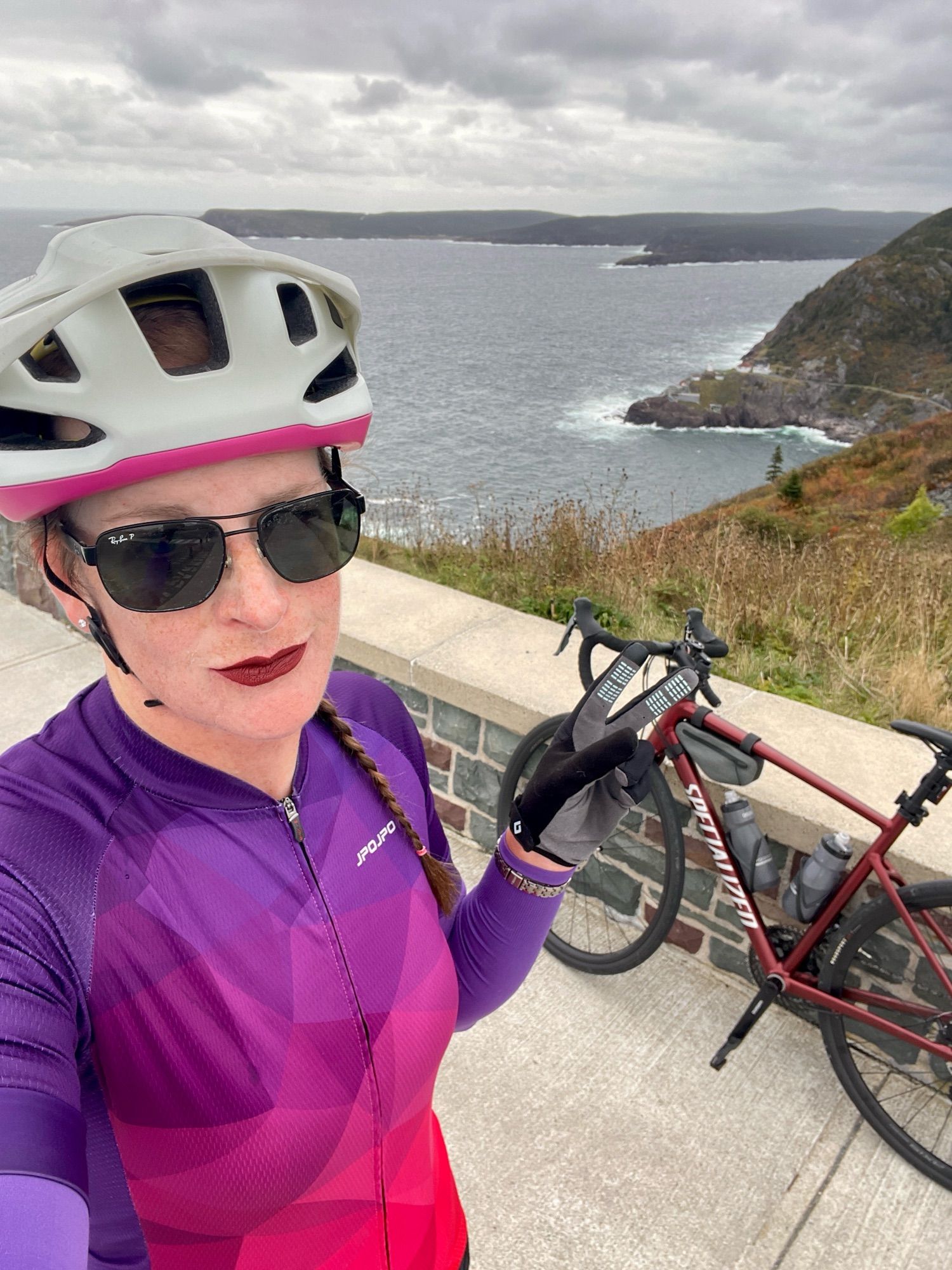 Cailín is standing on top of Signal Hill. Behind her is the Atlantic Ocean, Cape Spear, Fort Amherst and her maroon Specialized Allez road bike. She’s wearing a white and fuchsia cycling helmet, black aviator-style sunglasses, burgundy lipstick, a purple to red gradient cycling jersey, purple arm warmers, and black and grey full finger cycling gloves.