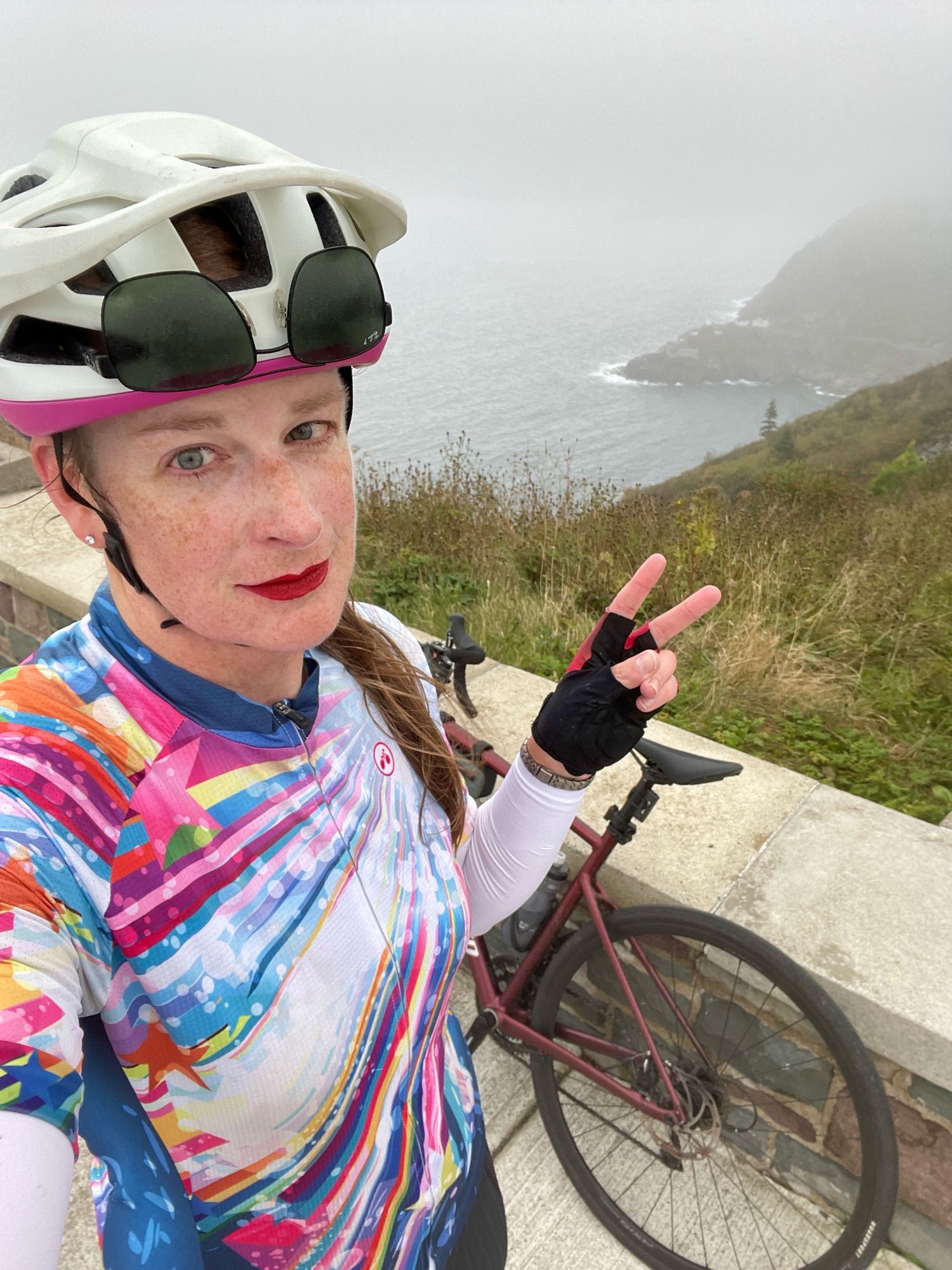 Cailín is standing at the top of Signal Hill. Behind her in the fog is the Atlantic Ocean, Fort Amherst, and her maroon Specialized Allez road bike. She’s wearing a white and fuchsia cycling helmet with her sunglasses stowed upside down in the front vents, red lipstick, a white and blue cycling jersey with colourful shooting stars on it, and red and black cycling gloves.