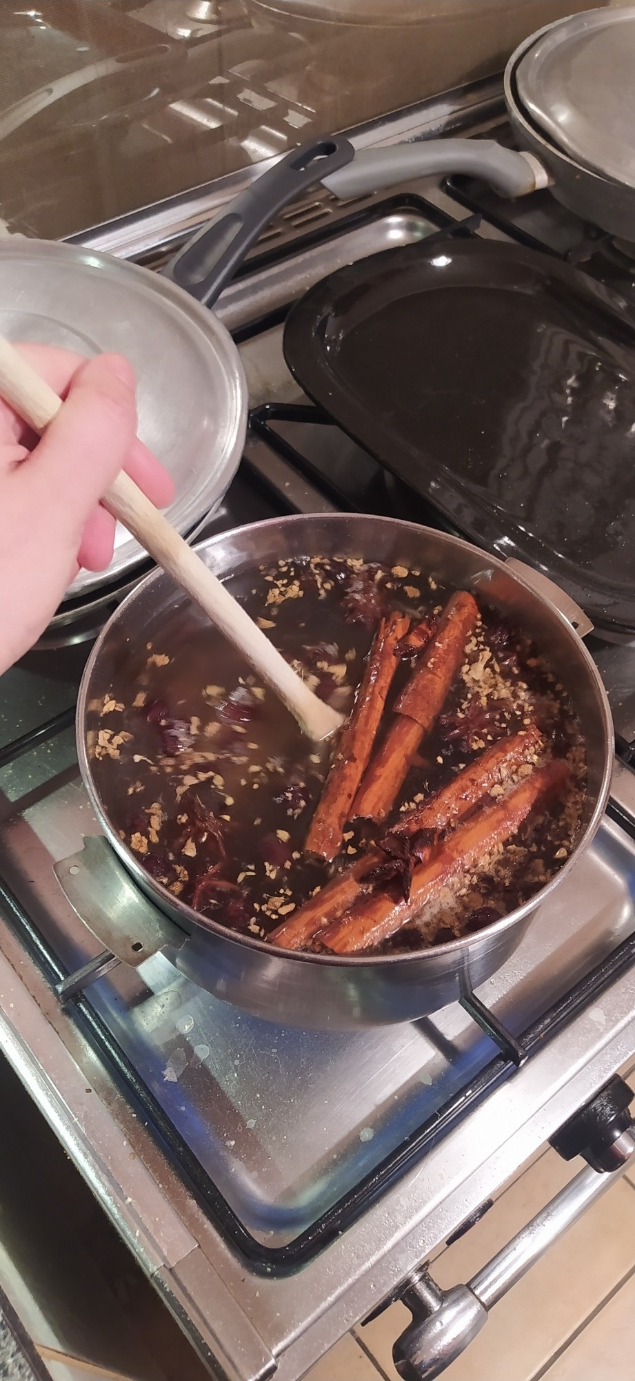 It's a pot full of water and ingredients such as cinnamon bars, ginger, lingonberries, star anise, cloves and sugar, with my hand holding a wooden spoon stirring it all, in the making of homemade finnish beverage named glögi