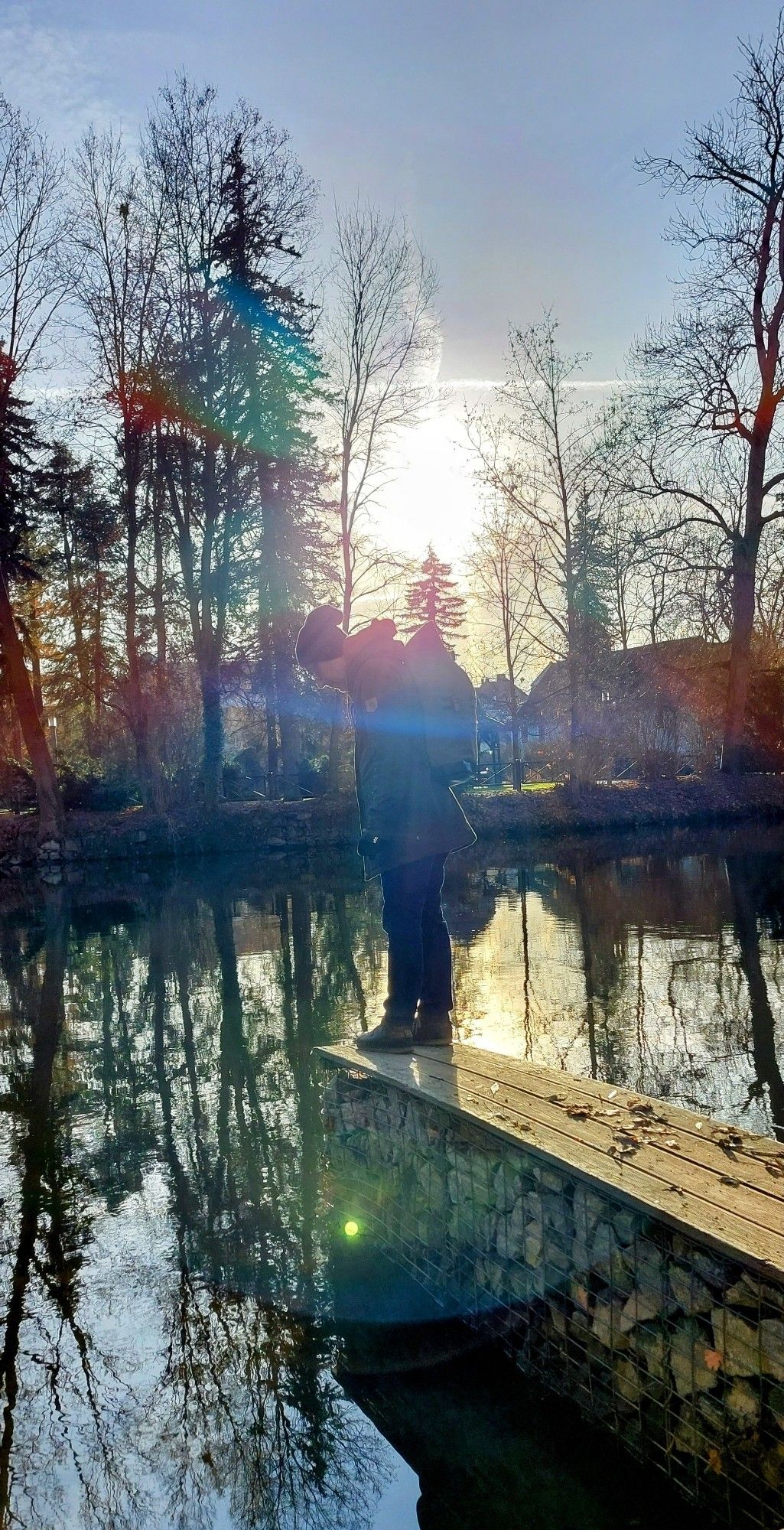 Man sieht einen Mann im Gegenlicht auf einem gemauerten Steg stehen. Im Wasser spiegeln sich hohe Bäume.