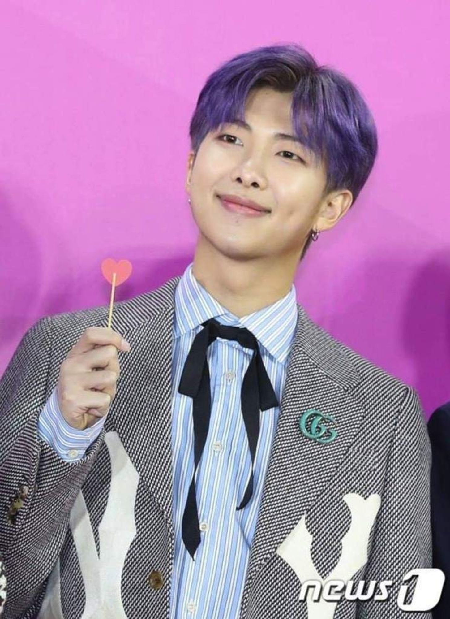 Namjoon is at a press event, photographed in front of a pinkish purple wall. He's holding up a heart shaped lollipop.