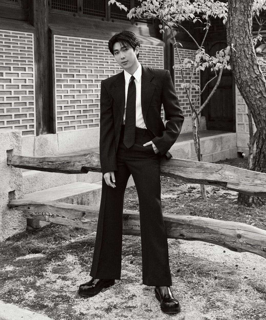 A black and white photo of Namjoon wearing suit and tie. He's standing outside in front of a brick building and a tree.