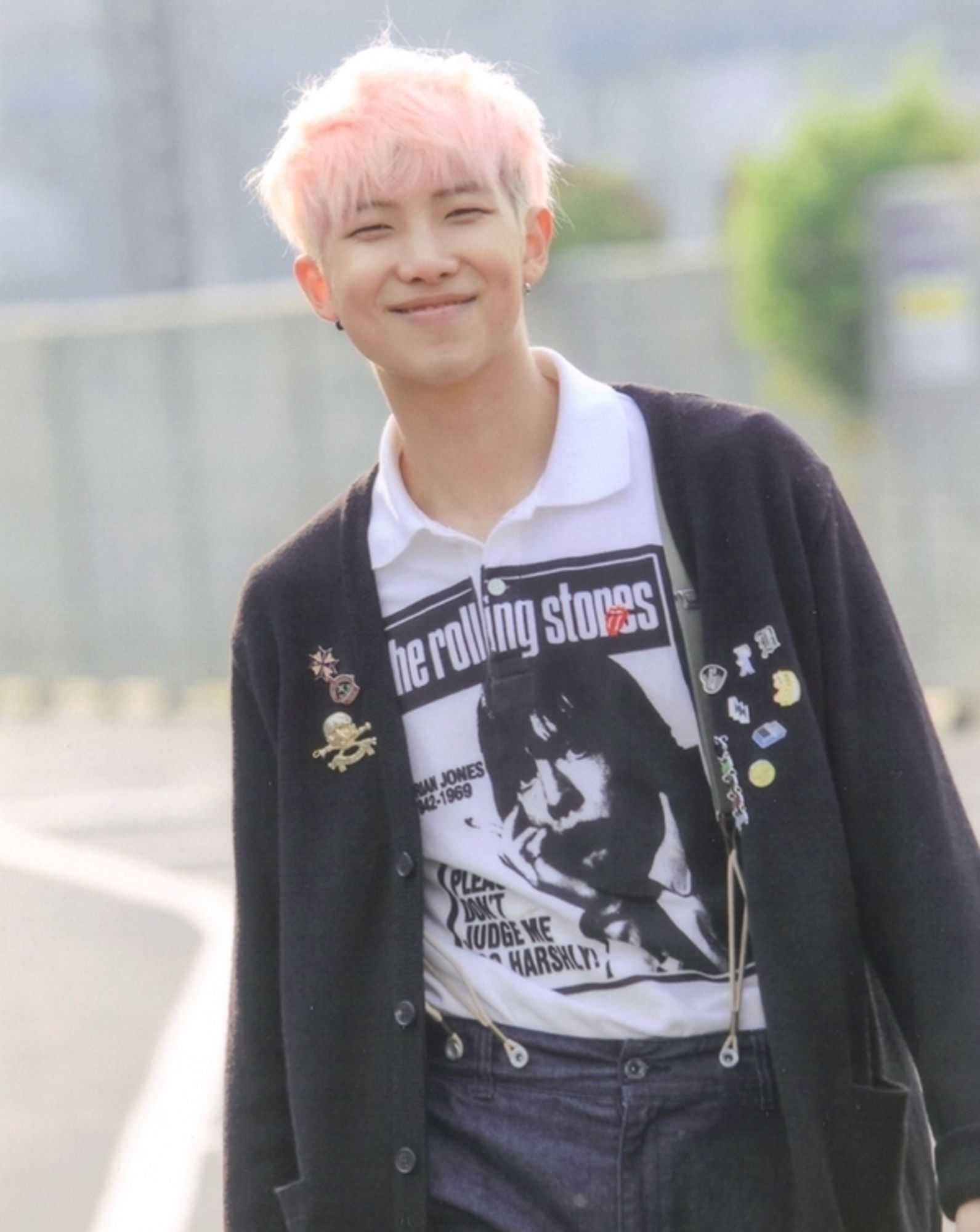 Namjoon has pink hair and is wearing a Rolling Stones t-shirt with a black cardigan with a bunch of pins on it. He's outside and smiling, walking toward the camera.