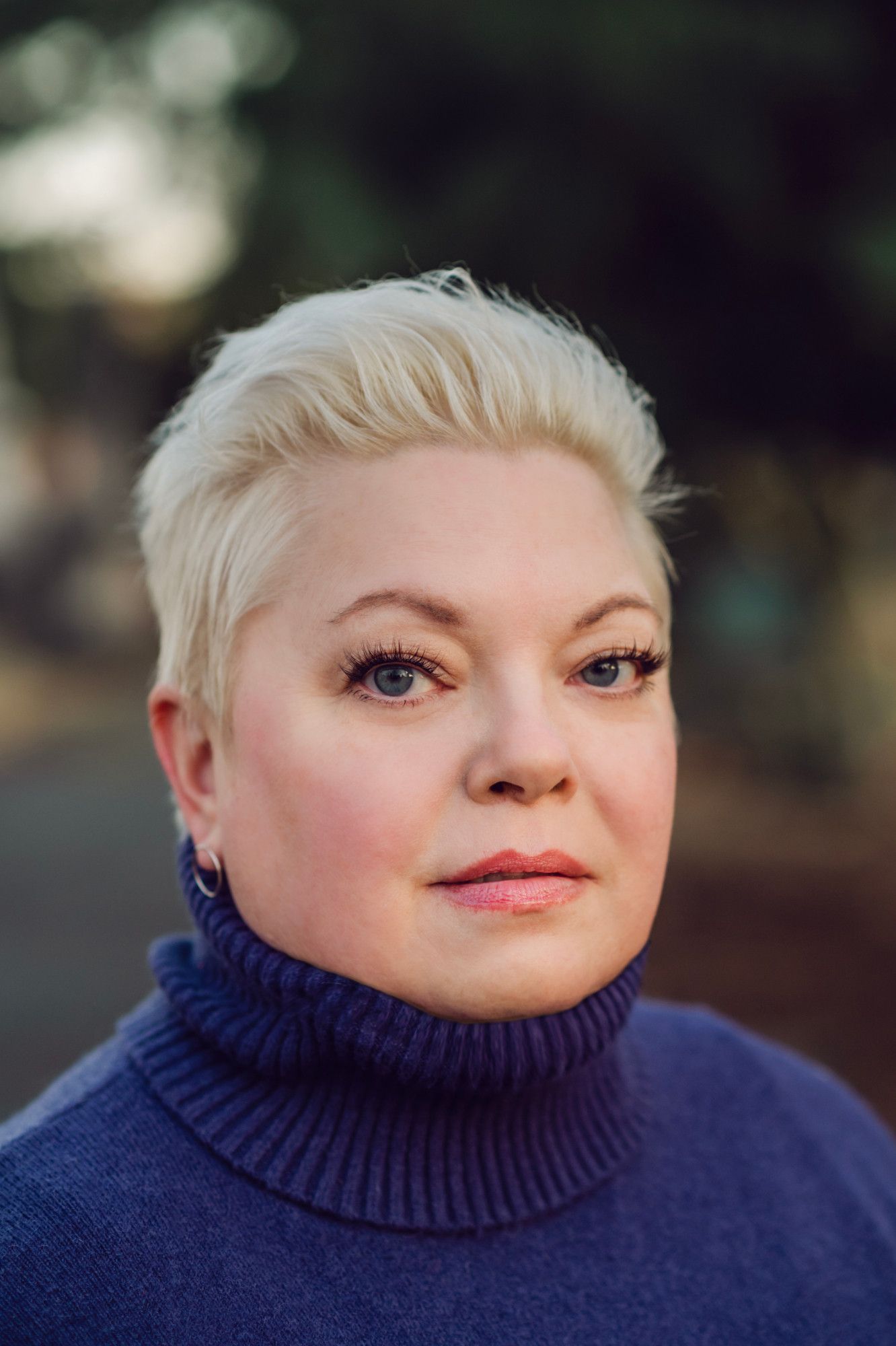 Head-and-shoulders color photo portrait or writer Nicole Wolverton, a fair-skinned woman with platinum blonde hair worn short and swept back from her forehead. She wears a ribbed turtleneck sweater in a dark, soft blue; small silver hoop earrings; and subtle makeup with long, dark eyelashes. She looks directly into the camera, smiling faintly.