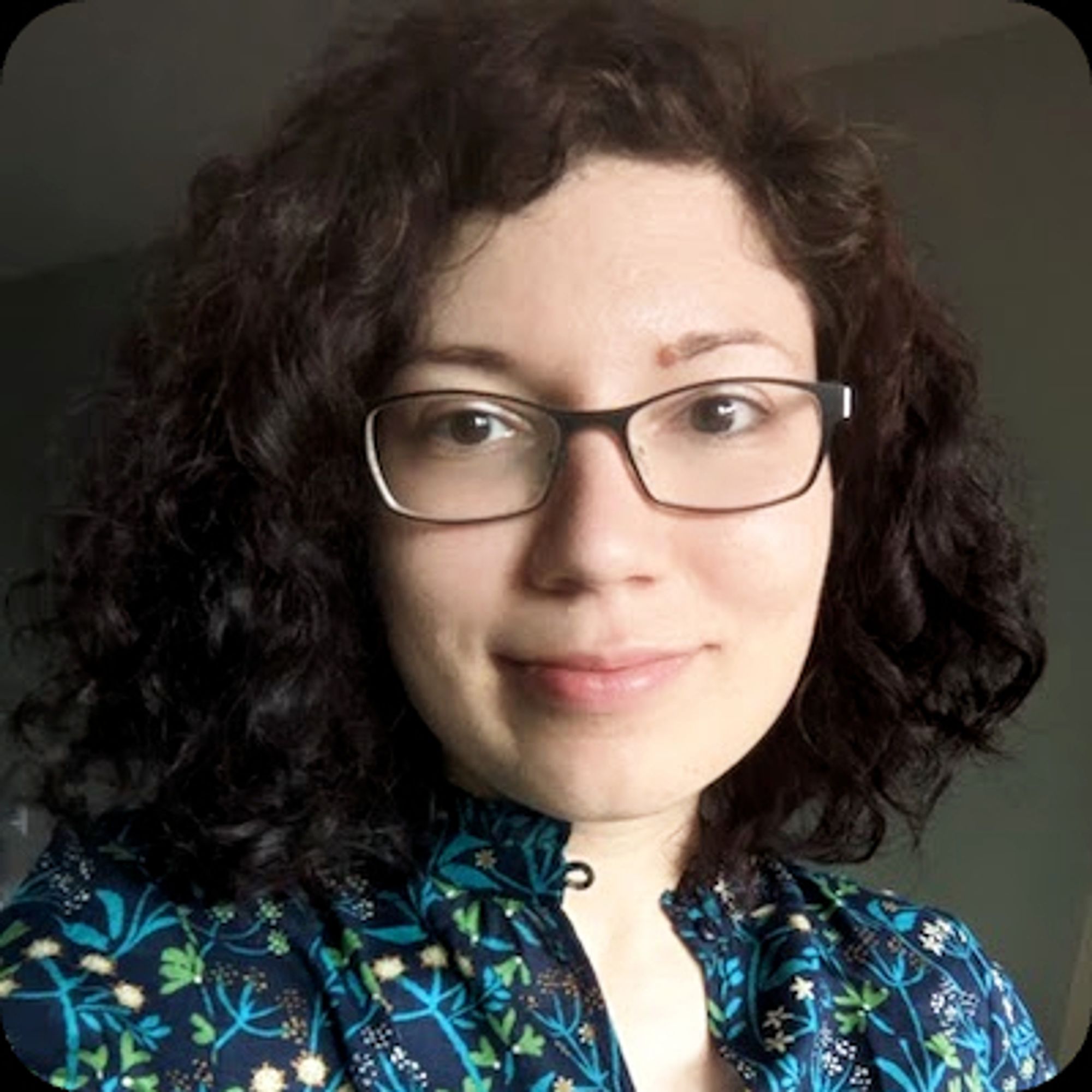 Author Diana Dima, a fair-skinned woman with brown eyes and curly dark-brown hair worn at shoulder length. She wears metal-framed glasses, a button-up shirt in a bold blue and green botanical print, and a little smile.