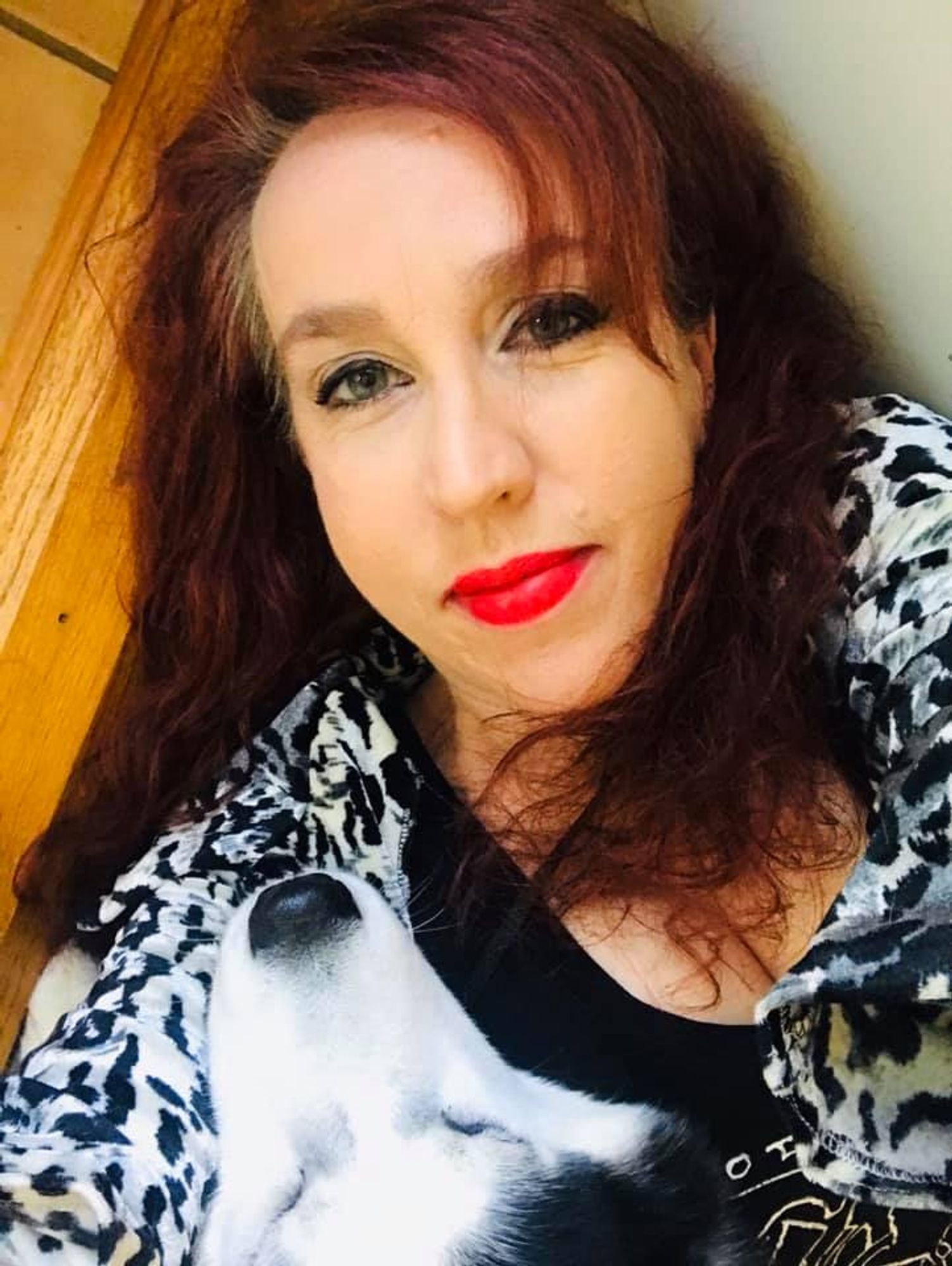 Author Anya Martin, a fair-skinned woman with gray-green eyes and long, burgundy hair. She wears a black-and-white animal print shirt, brilliant red lipstick, and sharp cat-eye winged eyeliner. She looks into the camera with a little smile while snuggling a large dog who obviously adores her.