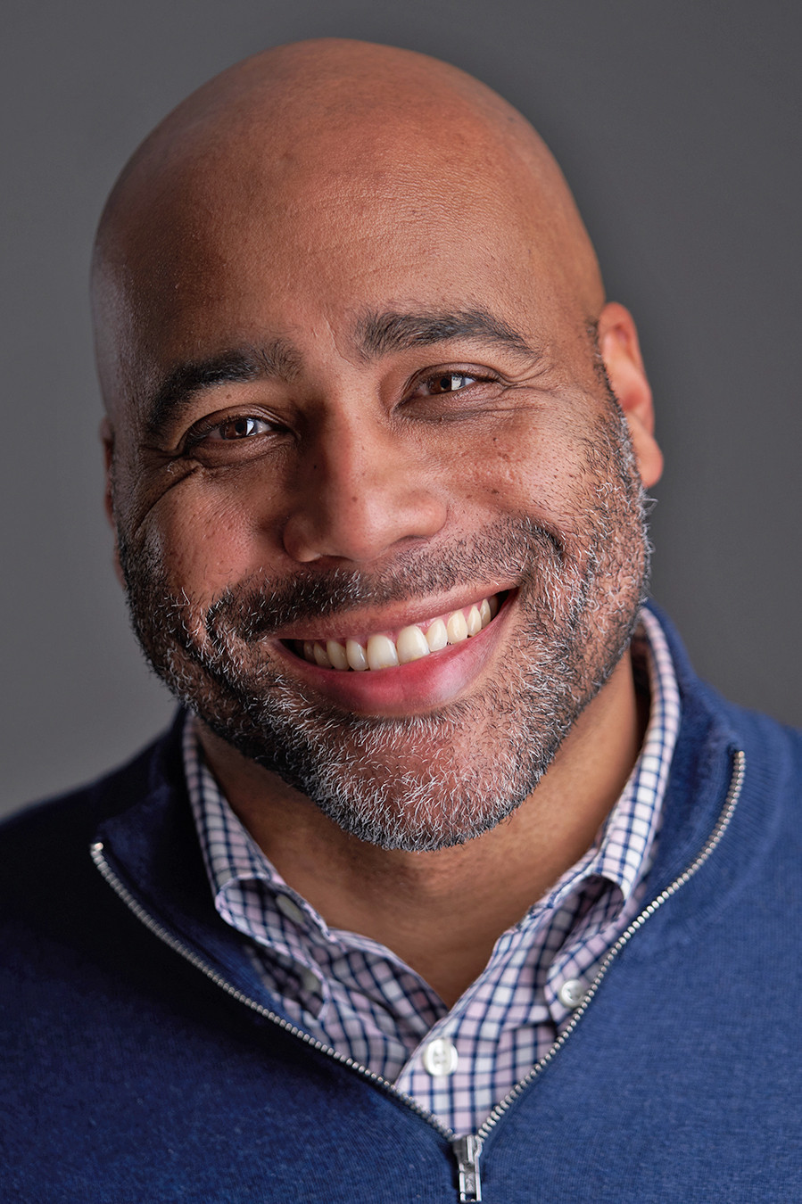 Author Johnny Compton, a Black man with a shaved head and short salt-and-pepper beard. He wears a windowpane plaid buttonup with a blue zippered cardigan. He smiles warmly into the camera.