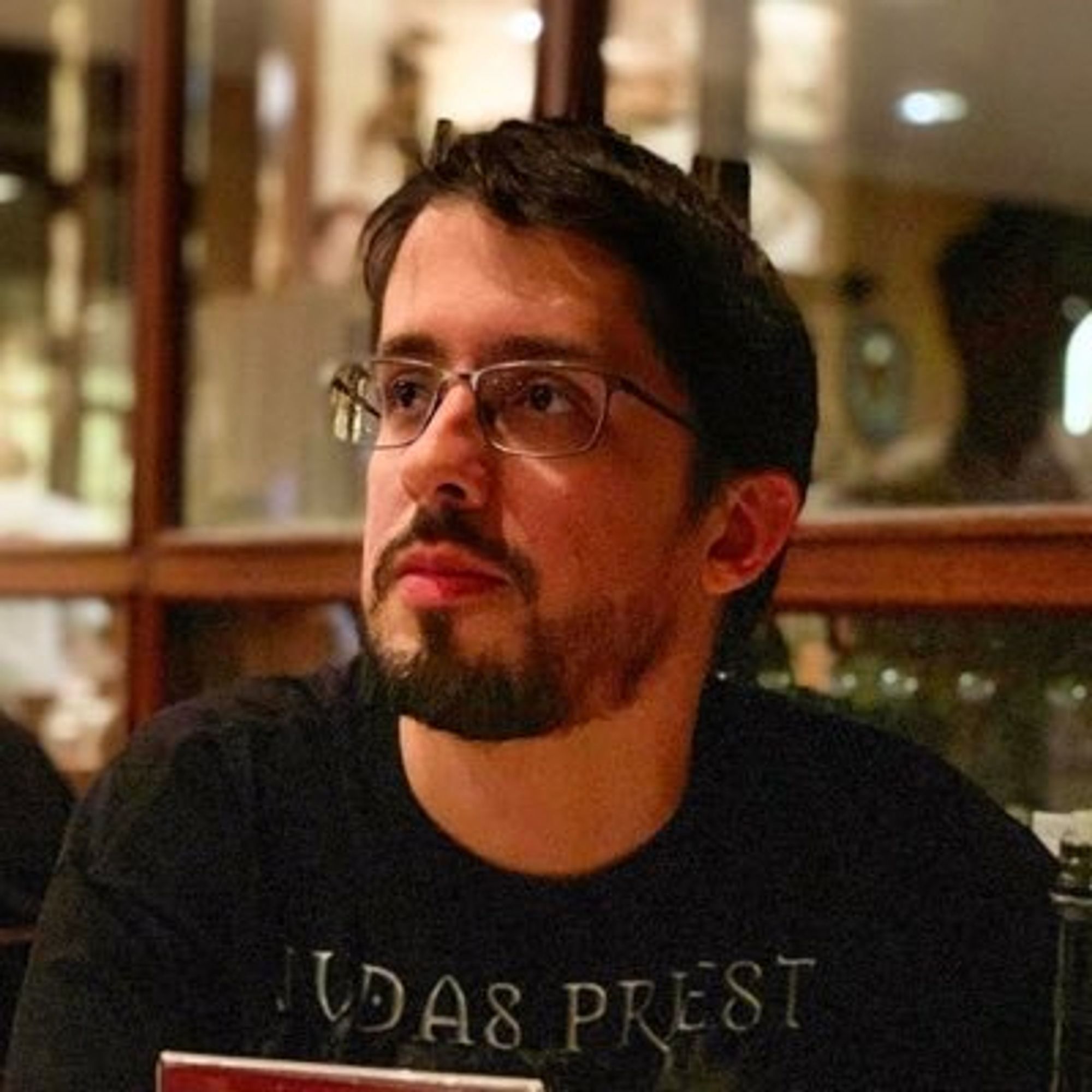 Casual color head-and-shoulders photo of writer Renan Bernardo, a man with dark eyes, straight dark hair, and a neatly trimmed dark beard and mustache. He wears glasses and a black Judas Priest shirt. He appears to be sitting in front of a many-paned window and is looking at something in the distance.