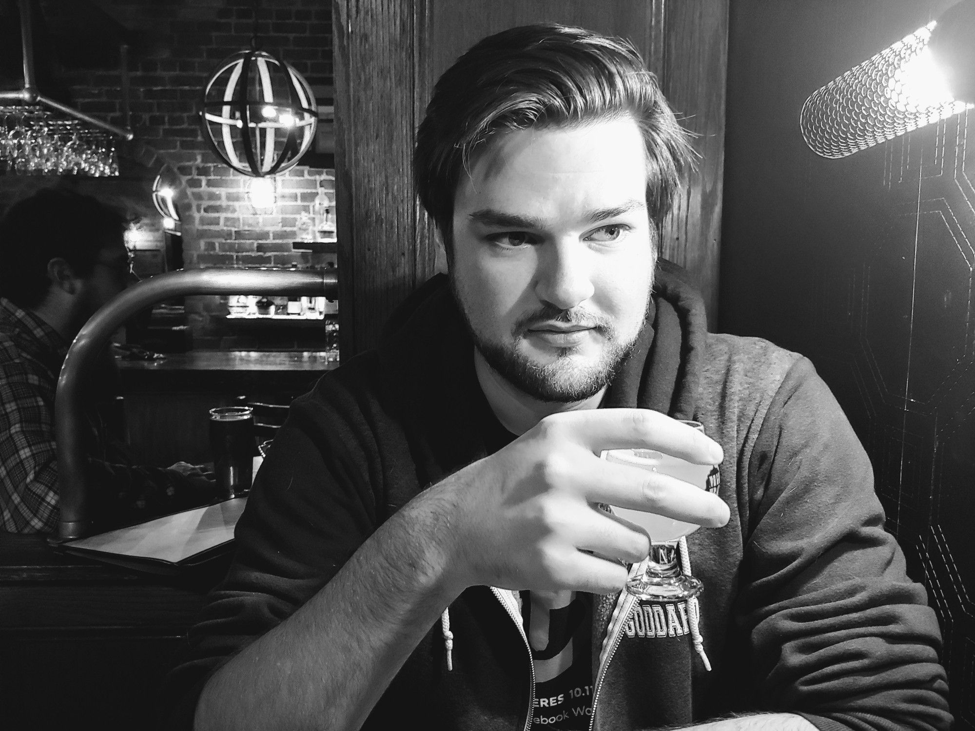 Black-and-white photo of author Sam Rebelein, a fair-skinned man with dark eyes, dark hair worn medium-short and partied on the left, and a short, well-kept beard. He sits in what looks like a bar, holding a cup and looking to the side with a contemplative expression. He is definitely up to something.