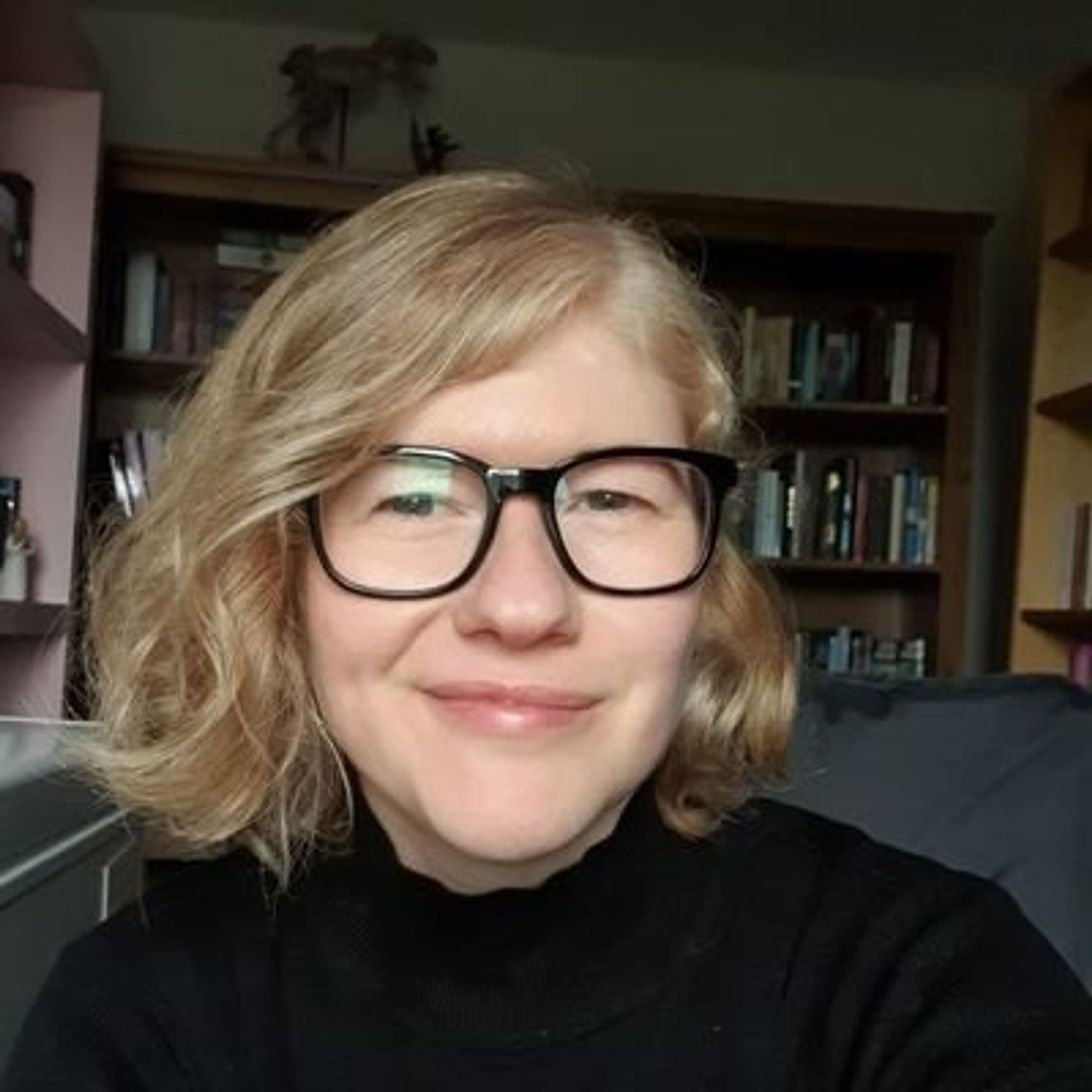 Color photo of Lindz McLeod, a fair-skinned woman with light blonde hair worn in a bob parted on her left. She wears a black mock turtleneck and glasses with large black frames. She sits in a room full of books and smiles craftily into the camera.