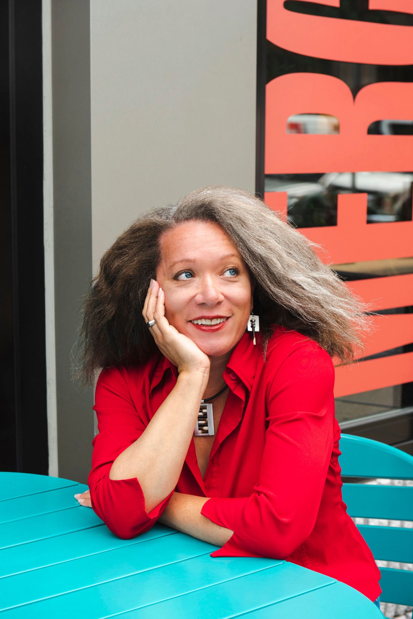 Color photo of author Tara Campbell, a Black woman with blue-green eyes, fair skin, and dark brown hair streaked with silver. She wears a red blouse and chunky silver jewelry. She sits at an outdoor table painted teal, in front of a café window. She rests her chin on one hand as she looks up and to the side, smiling. Photo by Hillary Deane.