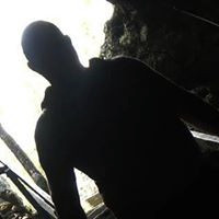 Waist-up photo of author Daniel Braum, a man silhouetted against a bright sky. Beyond the short cut of his hair, his features are impossible to see. Rocks loom behind him. He appears to be wearing a hoodie as he descends stairs with rough wooden banisters. He is going down, down into the depths.
