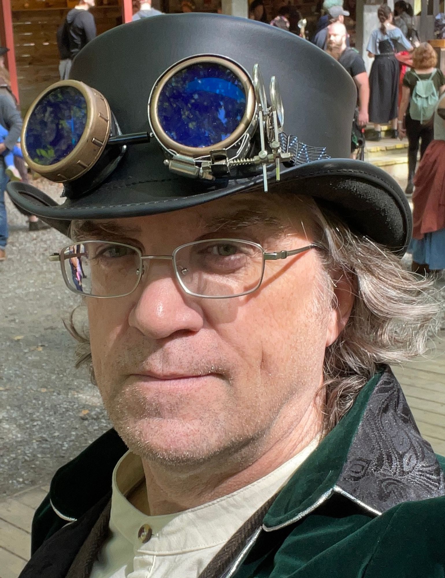 Casual color head-and-shoulders photo of Jeff Reynolds, a fair-skinned man with shoulder-length brown hair tingeds with gray. He wears metal-frame glasses, a brimmed hat with steampunk goggles, a banded-collar button-up shirt, and a green velvet coat (frock coat, perhaps?) with silver piping and a black leather collar. He appears to be standing on a boardwalk in a pedestrian area and looks at the camera with an alert expression.