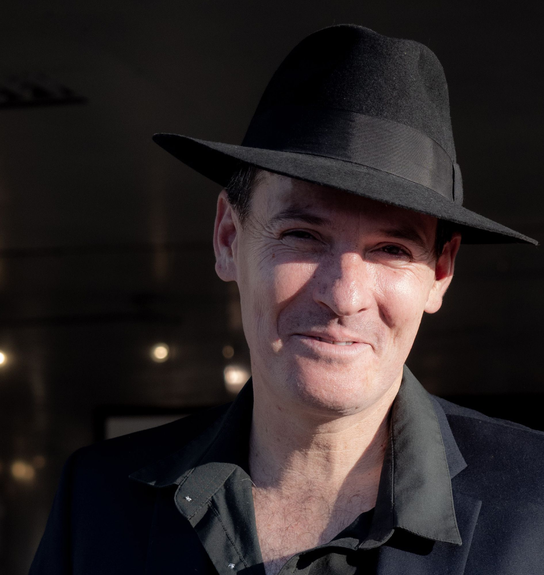 Color photo of author Jean-Paul Garnier, a thin, fair-skinned man with short, dark hair and dark eyes. He wears a black button-up and a black felt fedora. He looks into the camera and smiles as he squints in the bright sun. Photo by Kim Martin.