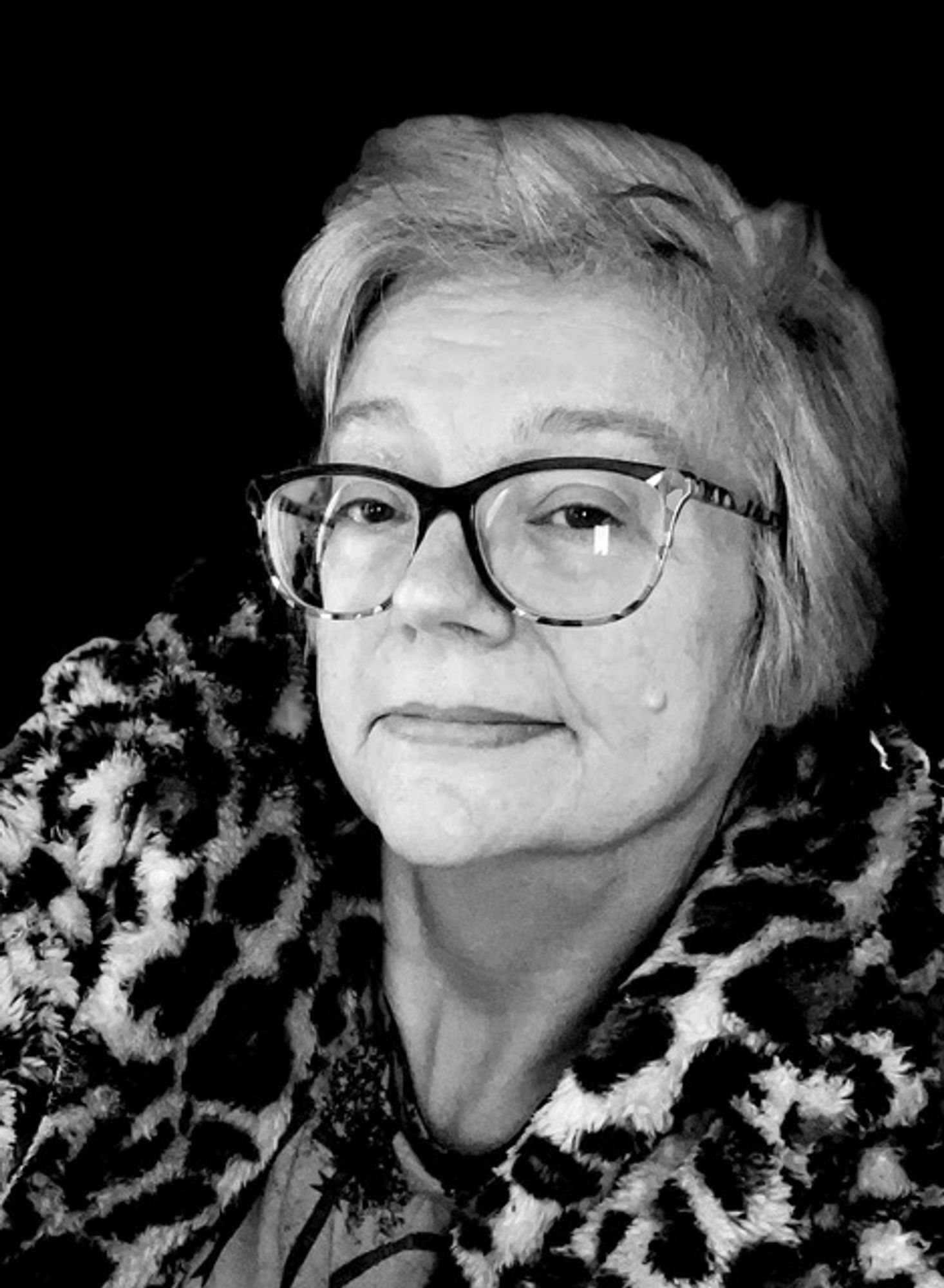 Black-and-white photo portrait of Cat Rambo, a fair-skinned person with short, light-colored hair. They wear a leopard-print faux fur, big glasses, and a patterned shirt. They look into the camera with a hint of a smile.