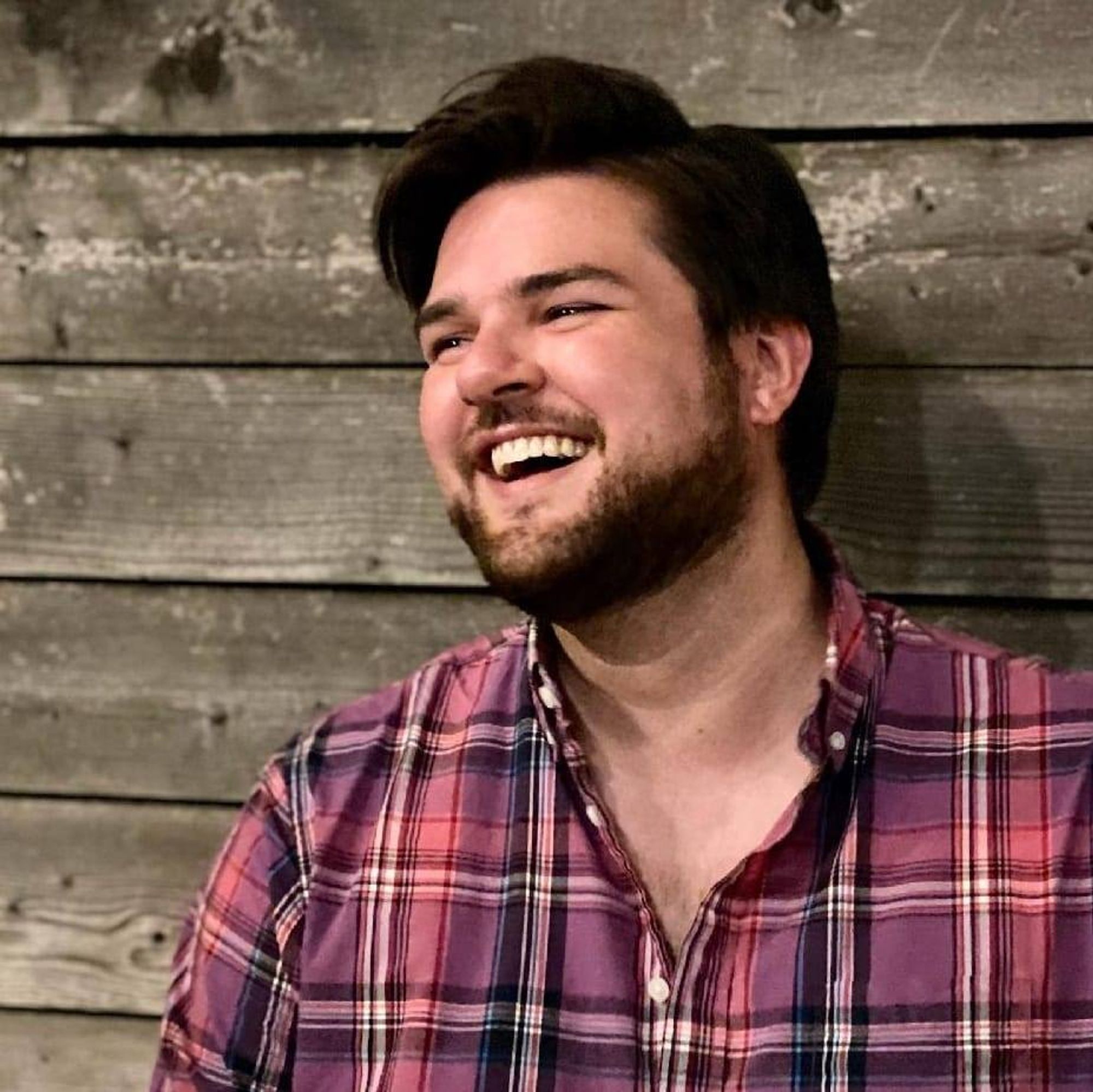 Color photo of author Sam Rebelein, a rosy-cheeked man with dark hair and beard. He wears a red plaid shirt and looks at something off-camera, laughing delightedly.