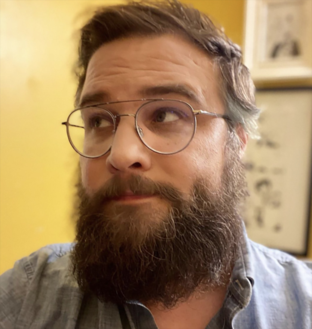 Close-up color photo of author Jordan Shiveley, a person with brown hair cut shortish and a full brown beard. They wear a collared denim shirt and a pair of aviator glasses and appear to be in some kind of interior, in front of a cartoon. They look up and to the side with a wary, bemused expression.