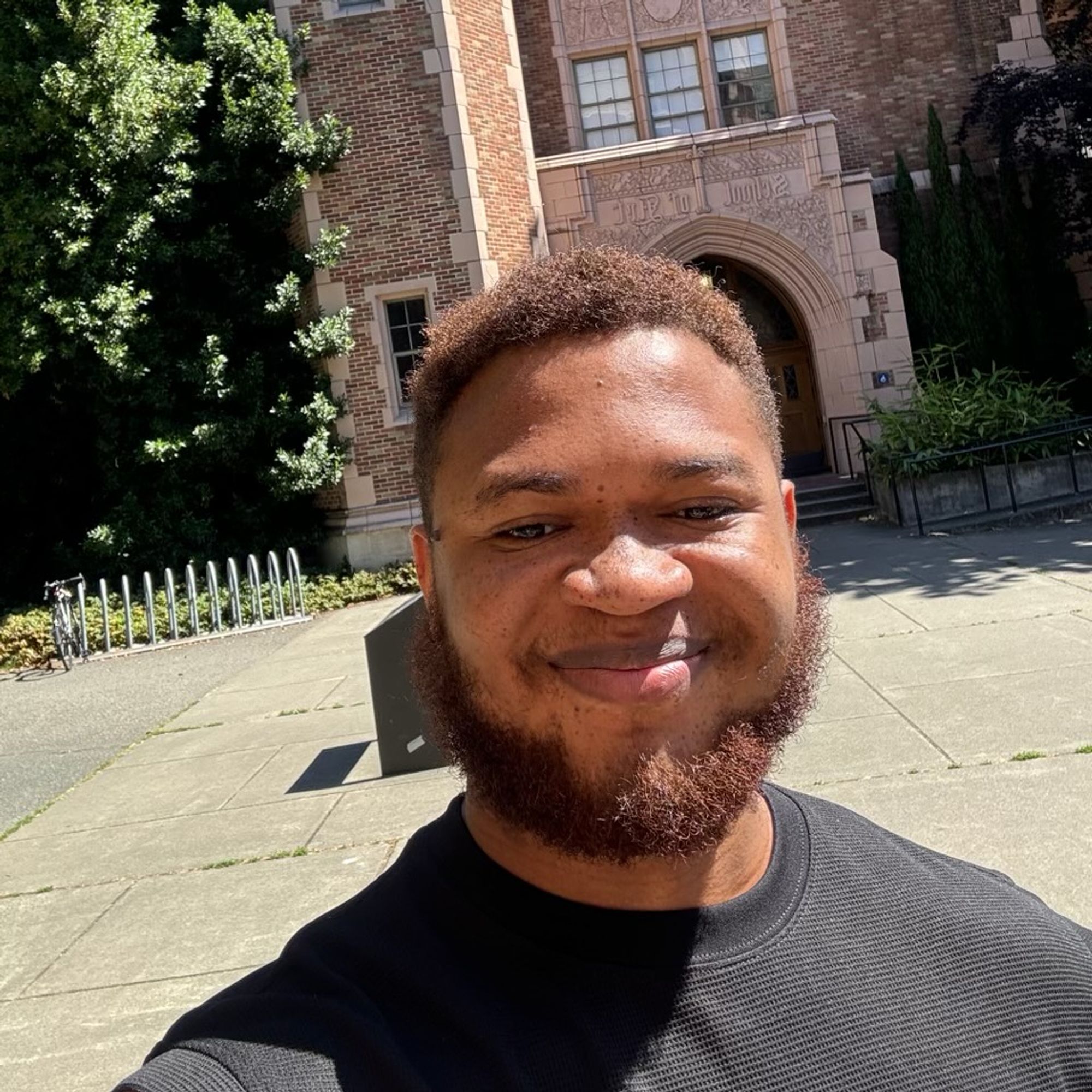 Color photo of Somto Ihezue, a Black man with freckled skin. He wears his curly red-brown hair short and has a matching beard. He wears a black waffle-weave shirt stands in front of a brick building that looks like a university building. He smiles warmly into the camera.