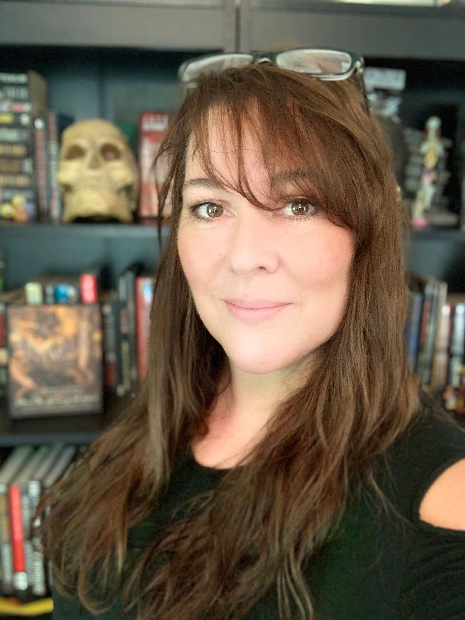 Head-and-shoulders color photo of Kelli Owen, a fair-skinned woman with brown eyes and long, chestnut brown hair with side bangs. She wears a pair of plastic-framed glasses on top of her head and a dark green cold-shoulder top. She stands in front of a bookcase that holds books and a human skull, smiling directly into the camera. Kelli does, anyway. It's hard to tell where the skull is looking.