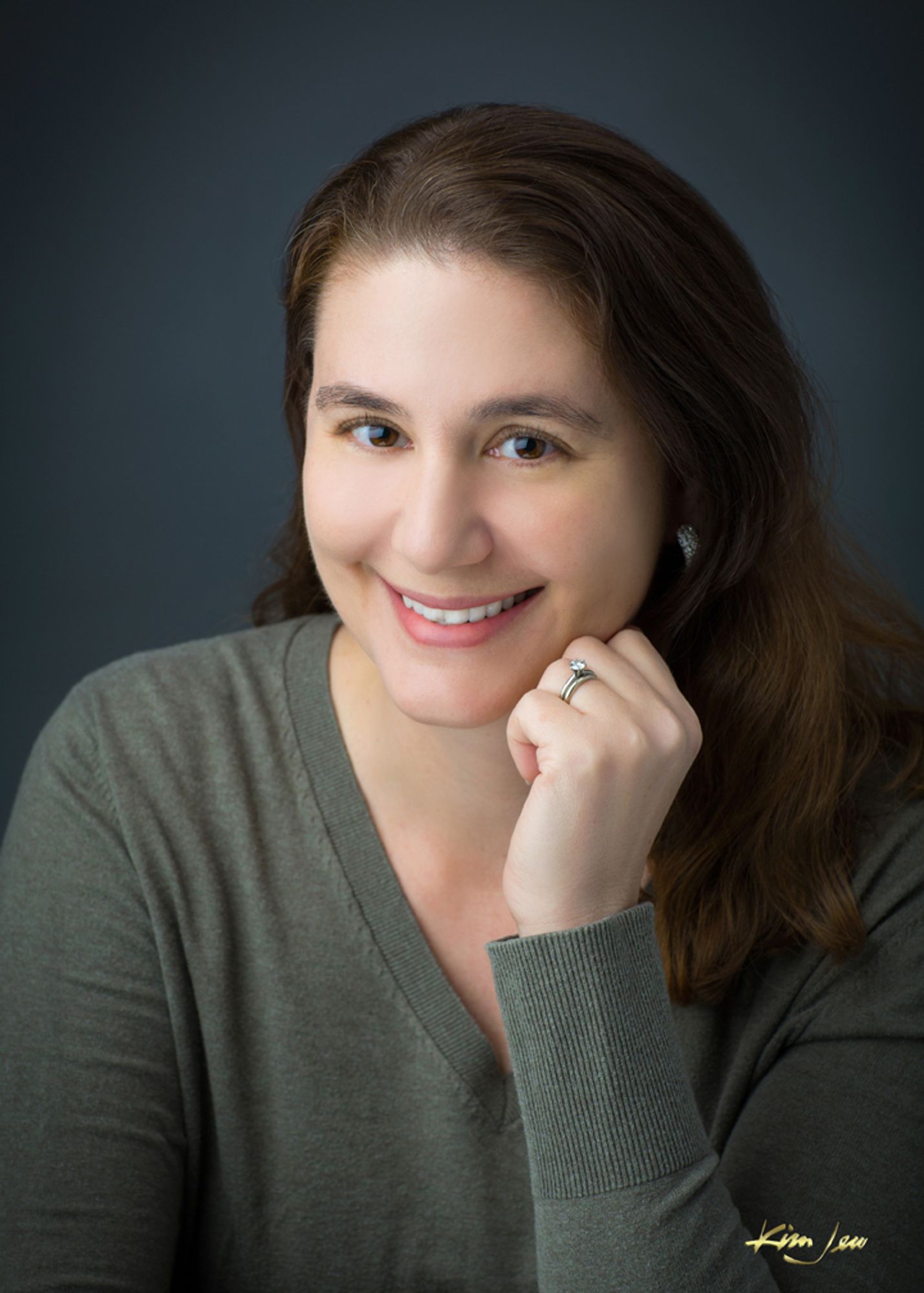 Color photo of Lauren C. Teffeau, a fair-skinned woman with red-brown hair worn long and without bangs. She wears a gray-green V-neck sweater, and she rests her chin on one hand as she smiles warmly at the camera.