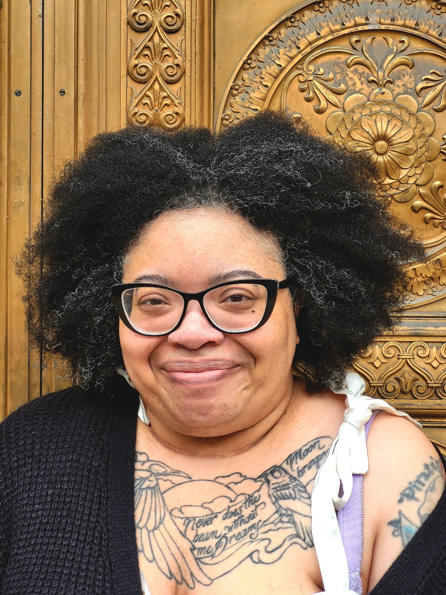 Color photo of author Donyae Coles, a Black woman with a luxuriant natural. She wears large cat-eye glasses with black frames, a bag on one shoulder, and a black cardigan. The cardigan is pulled off one shoulder, exposing elaborate tattoo work. She looks into the camera, smiling like she's just thought of a joke.