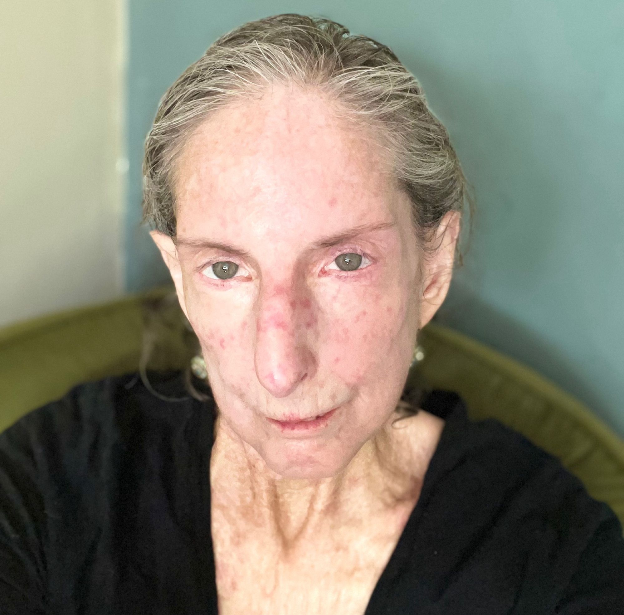 Color photo of Jessica Reisman, a fair-skinned person with graying brown hair worn pulled back and without bangs. They wear a black V-neck shirt and sit on a green upholstered chair. They look up at the camera with a small smile.