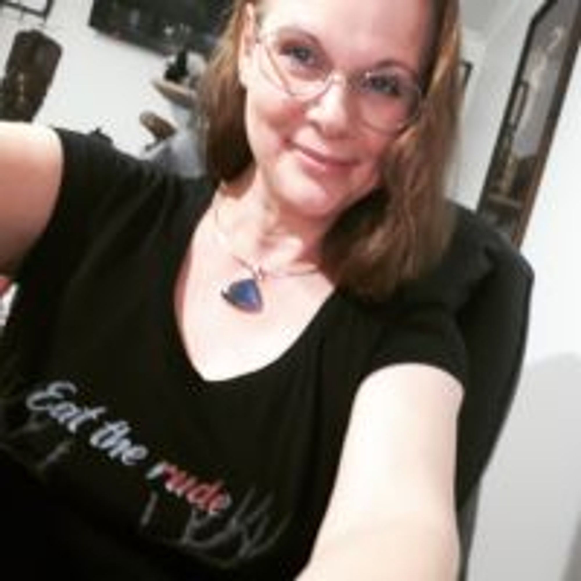 Blurry selfie of author L.E. Daniels, a fair-skinned woman with red-gold hair worn at shoulder length. She wears round glasses, a large pendant on a gold chain, and a black T-shirt that says "Eat the rude" in script. She smiles as if she has a secret, and her arms reach to the camera, as if they are holding it...or maybe she's coming out of your screen.