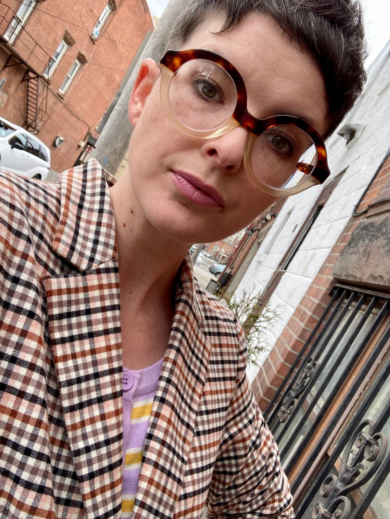 Author Dee Holloway, a fair-skinned woman with short, dark hair. She wears a striped pastel shirt; a checked blazer; and large, round, two-toned glasses. She regards the camera with the ghost of a smile.