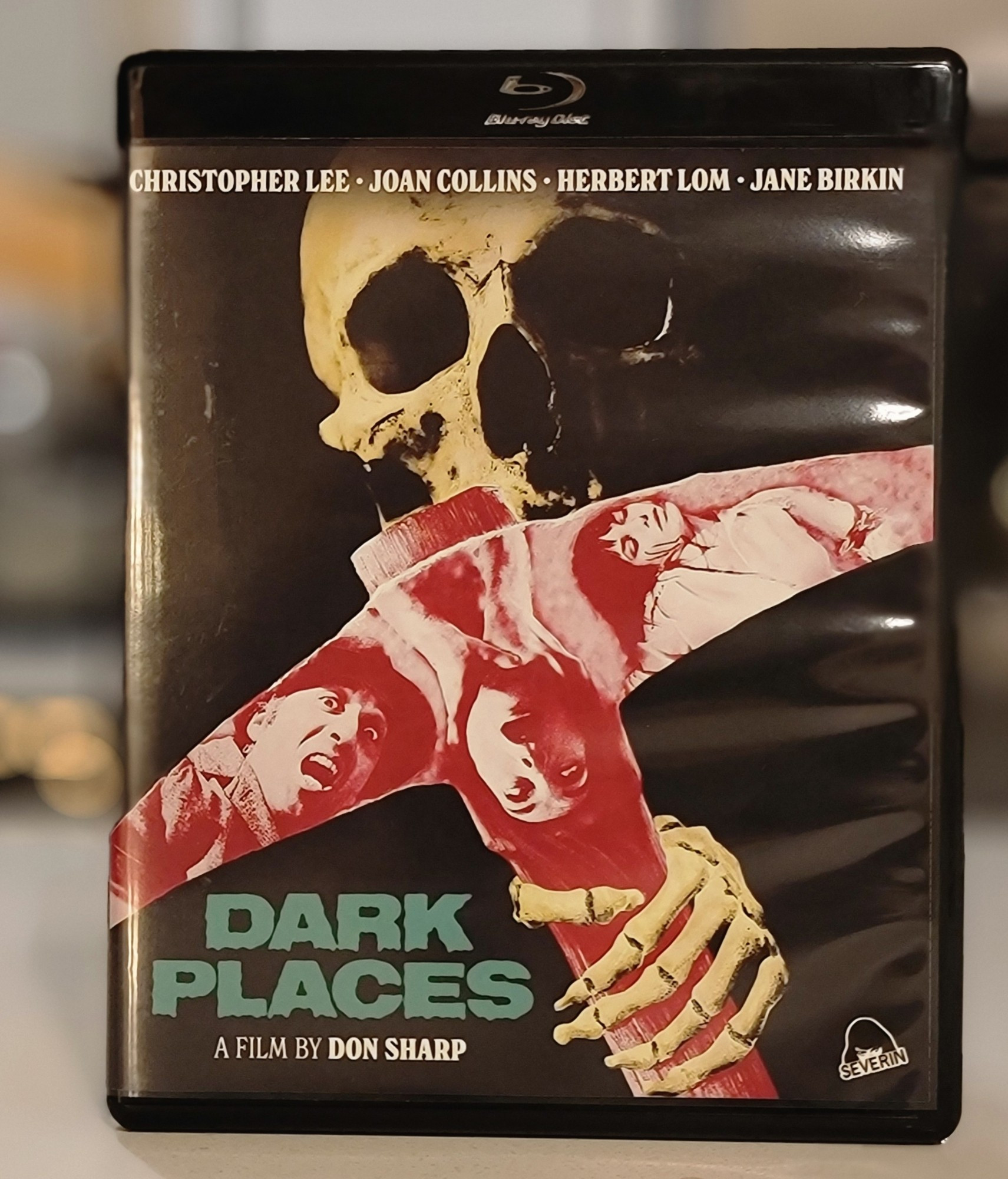 Blu ray cover for the film Dark Places. The image shows a skeleton with a pickaxe. There are images of various frightened or dead people pictured in the pickaxe image.